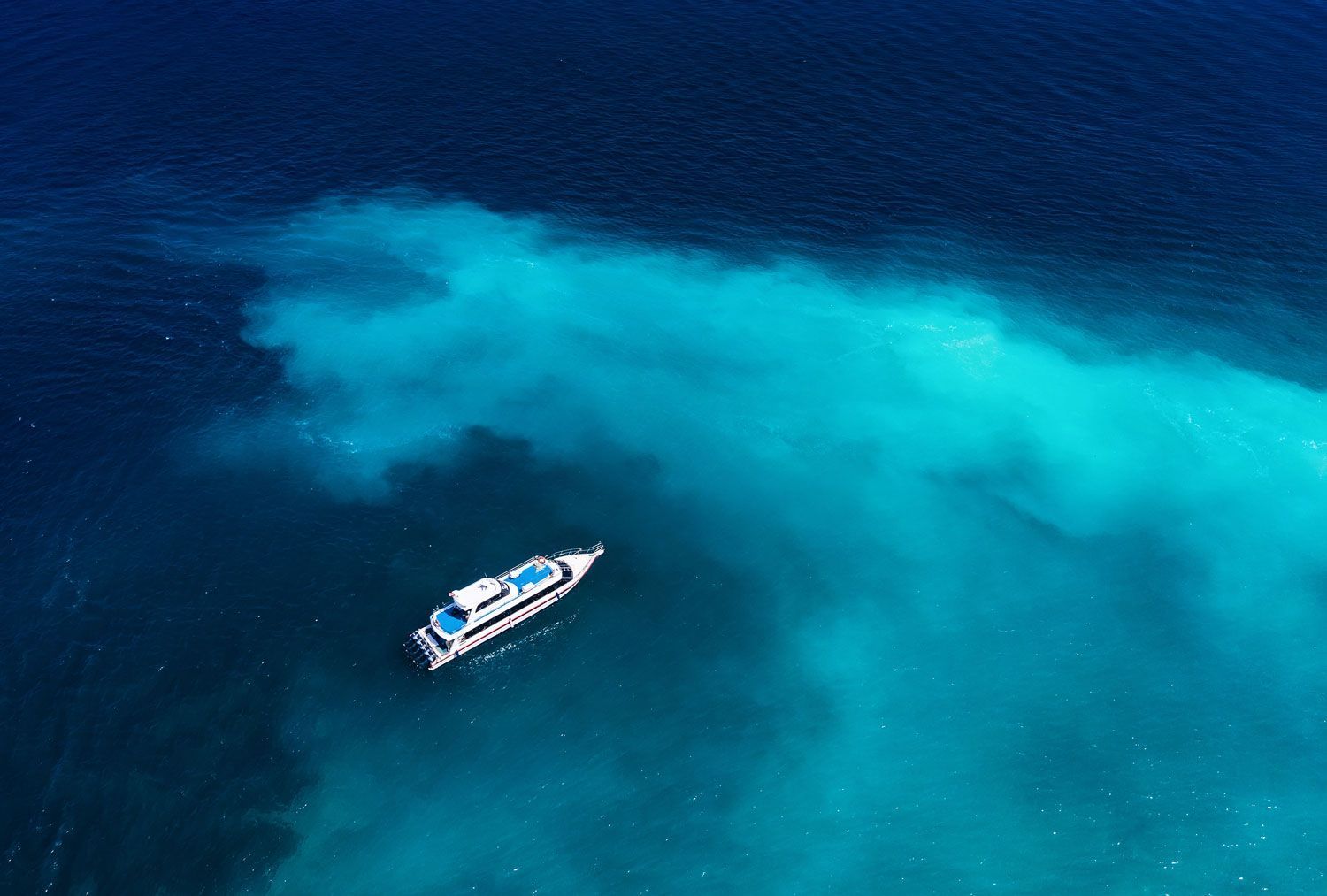 An aerial view of a white yacht floating on vibrant, clear turquoise water, surrounded by deeper blue ocean.