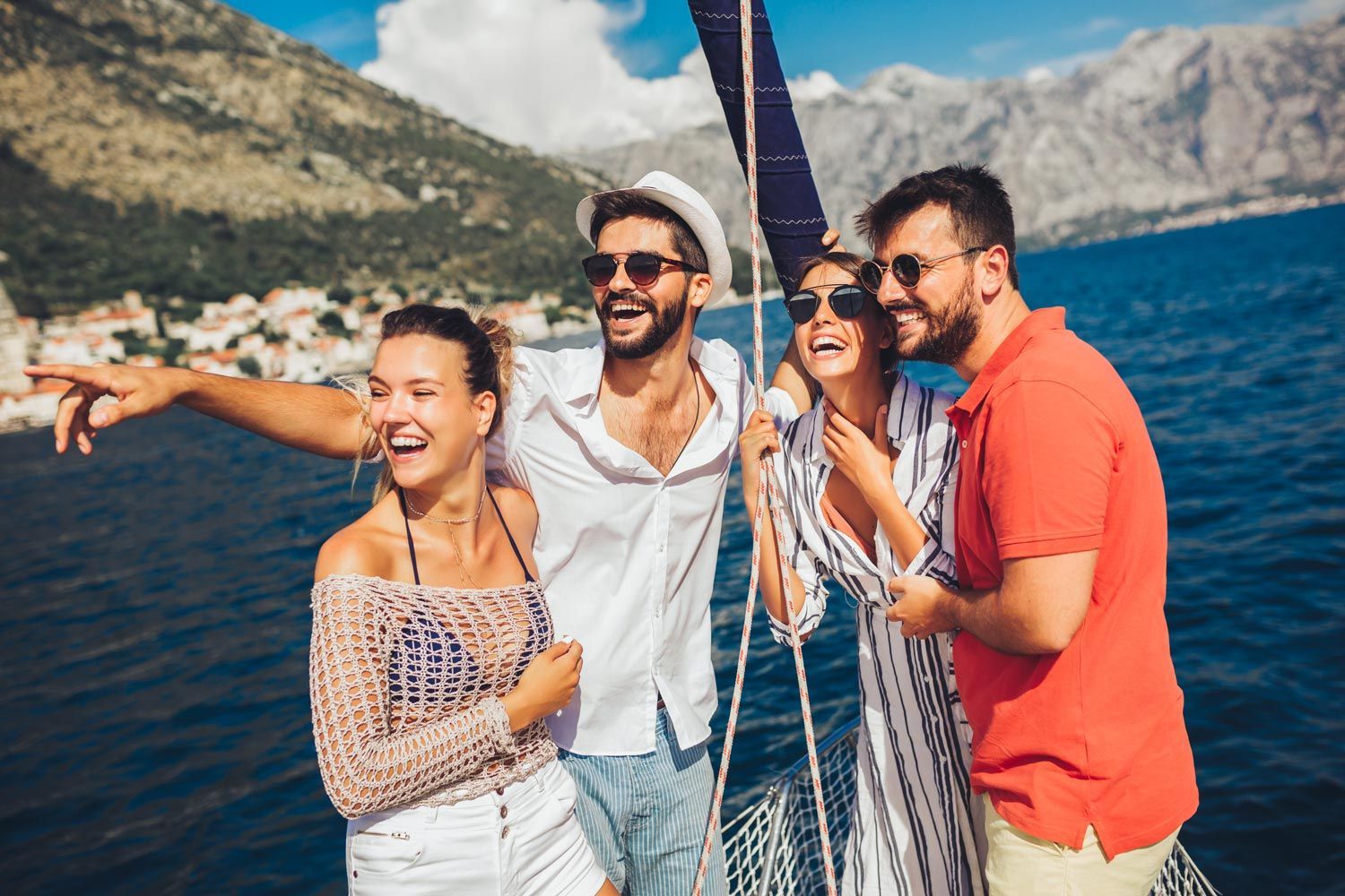 Four friends on a sailboat pointing toward a mountainous coastline, laughing under a sunny sky.