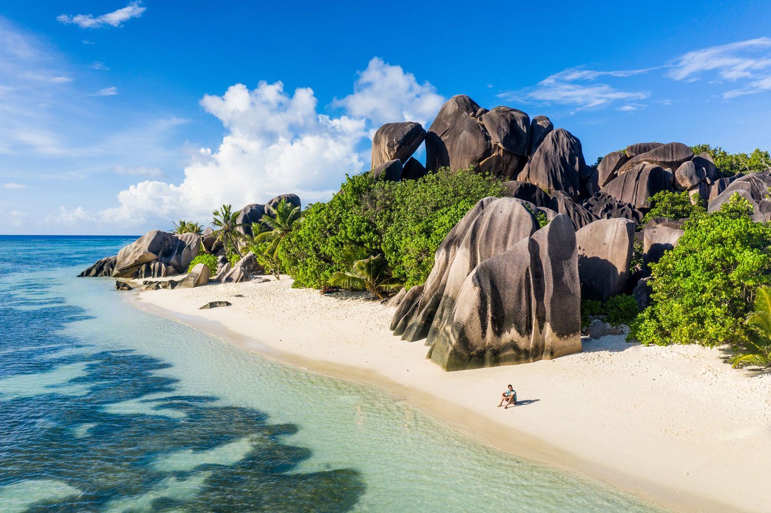 An aerial view of a white sandy beach with large granite boulders, lush greenery, and clear turquoise water.
