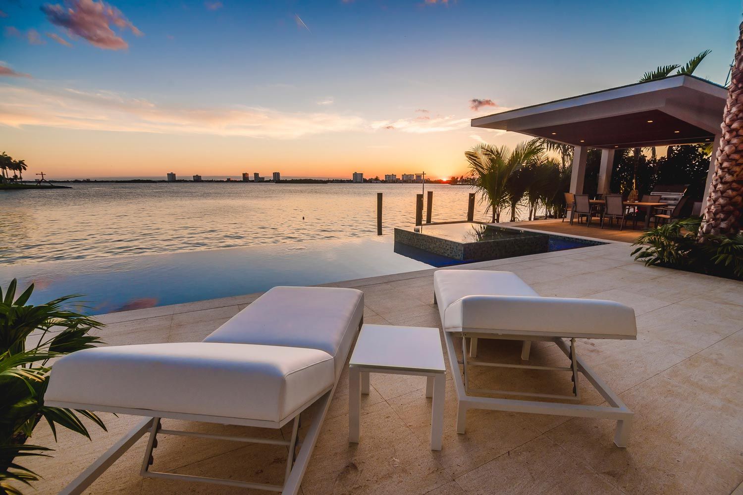 Two white lounge chairs on a stone patio overlooking an infinity pool and a calm bay at sunset with a nearby gazebo.