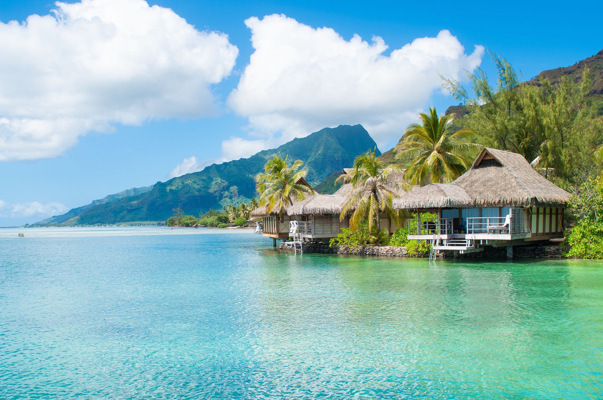 Overwater bungalows with thatched roofs sit in a turquoise lagoon against a backdrop of lush mountains and blue sky.