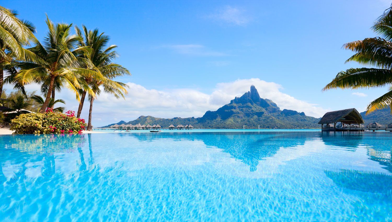 A clear blue infinity pool overlooks a tropical lagoon with overwater bungalows and a dramatic mountain peak.