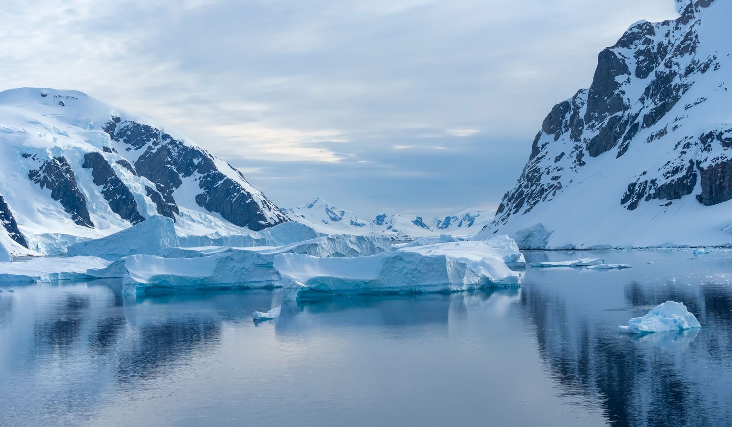 Snow-covered mountains flank a calm, dark waterway filled with blue-tinted icebergs under a pale, overcast sky.