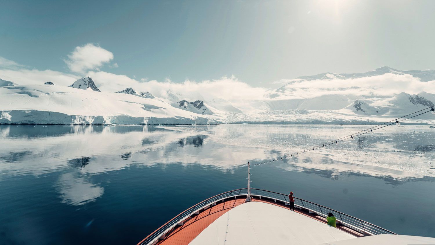 A view from the bow of a cruise ship moving through calm, icy waters toward snowy, sunlit mountains in Antarctica. A view from the bow of a cruise ship moving through calm, icy waters toward snowy, sunlit mountains in Antarctica.