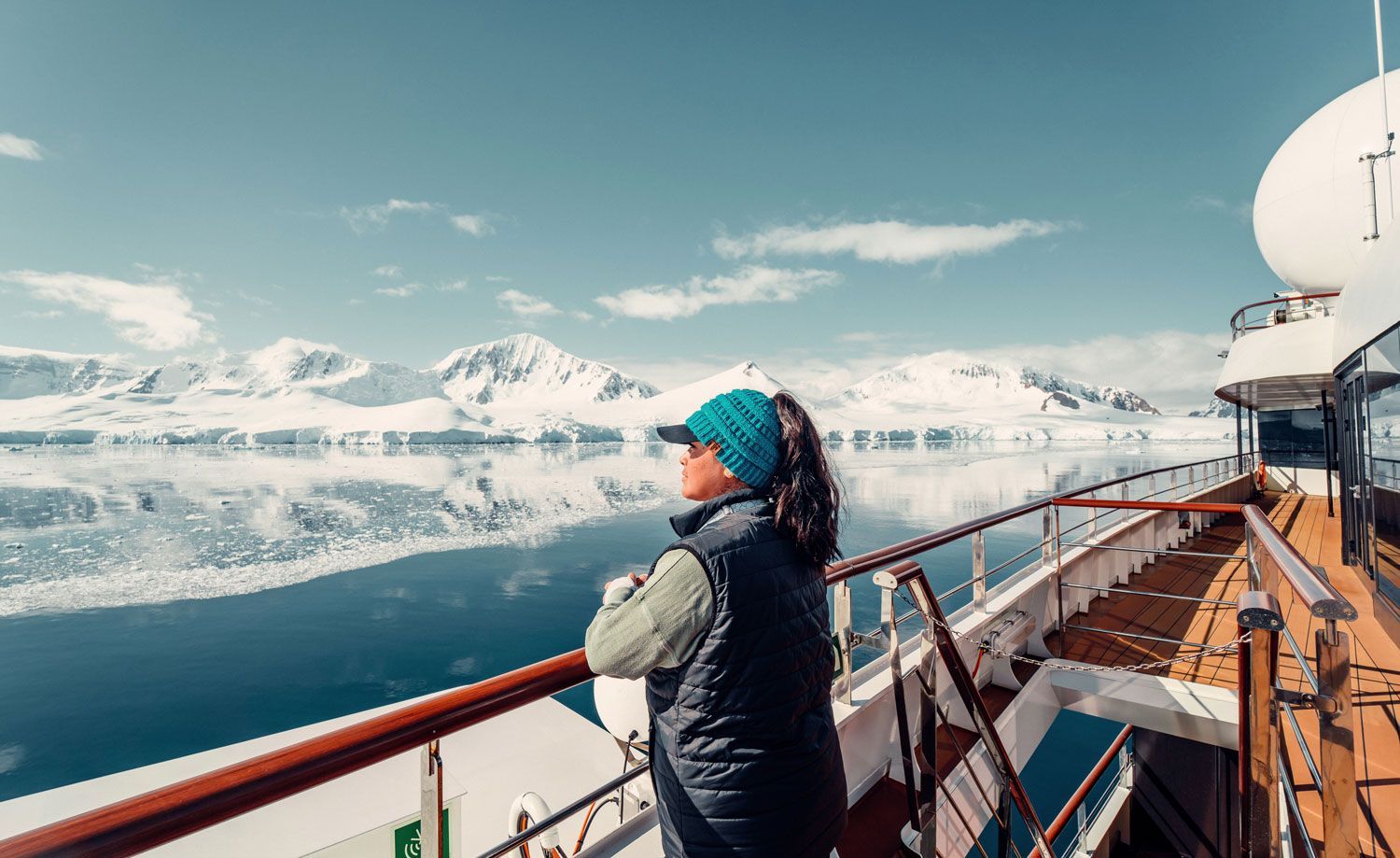 A person in a blue hat and dark vest stands on a ship deck looking out at a serene, snow-covered mountain landscape.