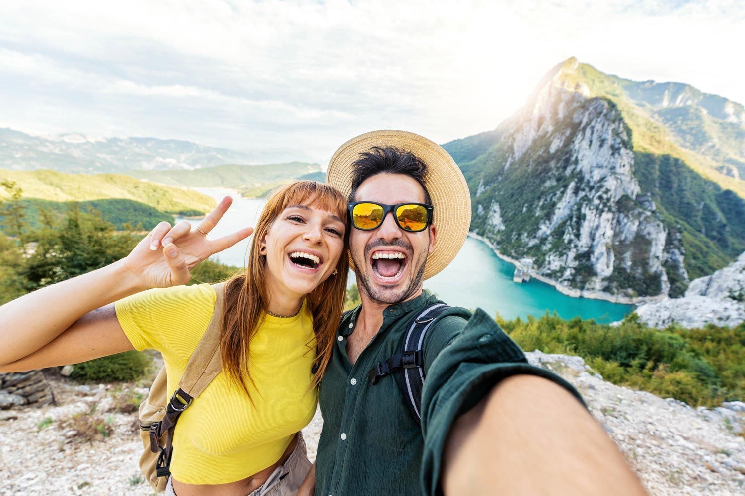 A joyful couple in a yellow shirt and green shirt takes a selfie overlooking a turquoise river and rocky mountains.
