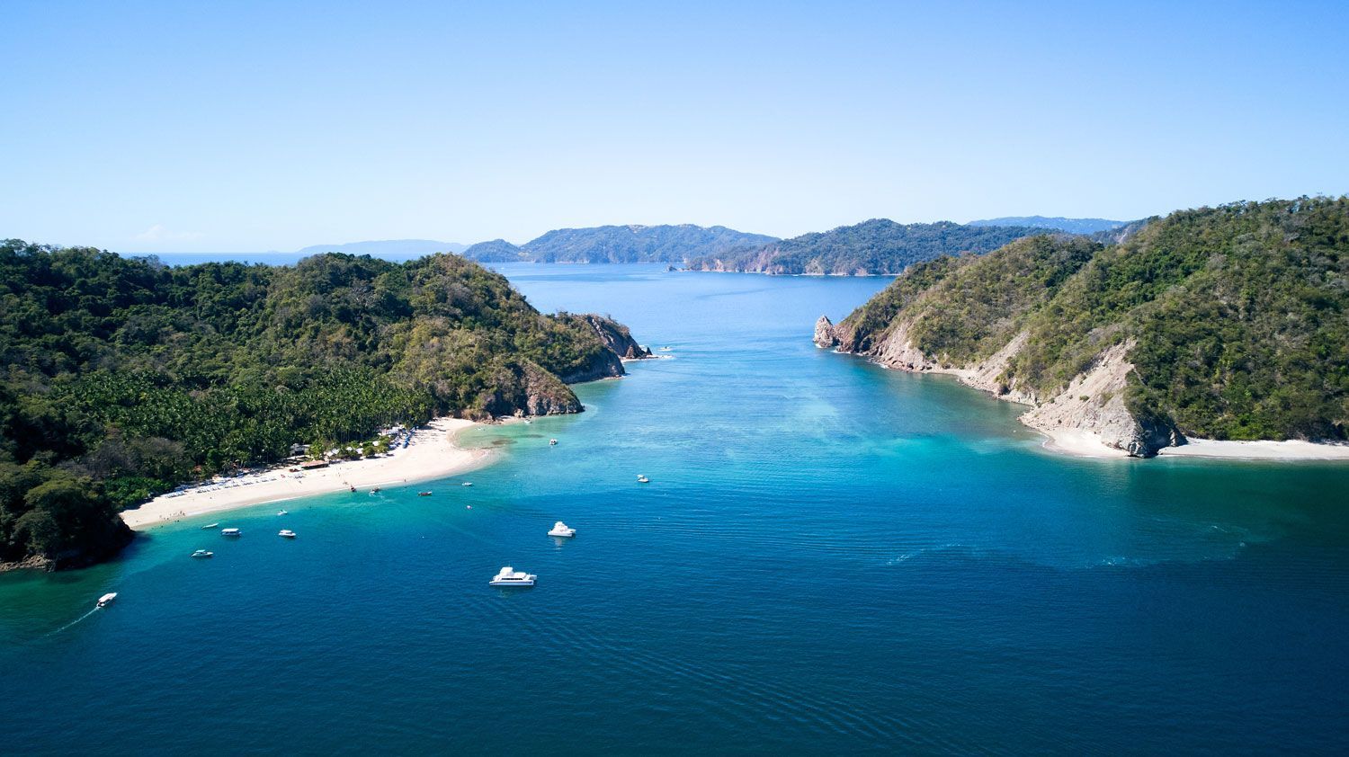 Aerial view of a blue tropical bay nestled between lush, forested hills with white sandy beaches and small anchored boats.