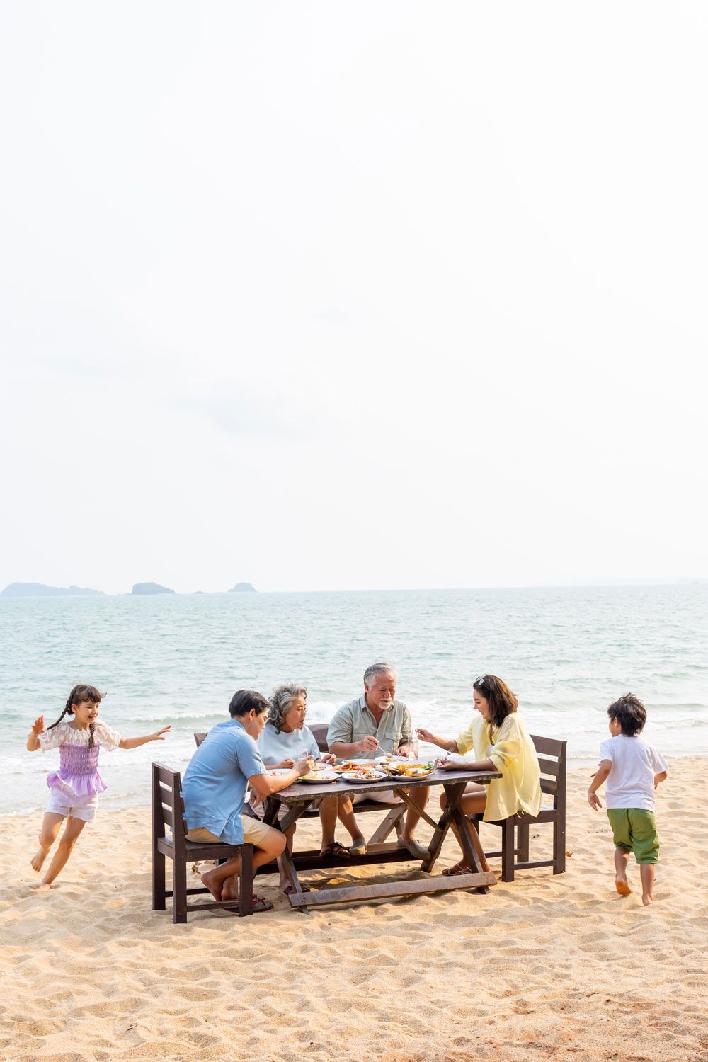 A family sits at a wooden table on a sandy beach eating, while two children play in the sand nearby. A family sits at a wooden table on a sandy beach eating, while two children play in the sand nearby.