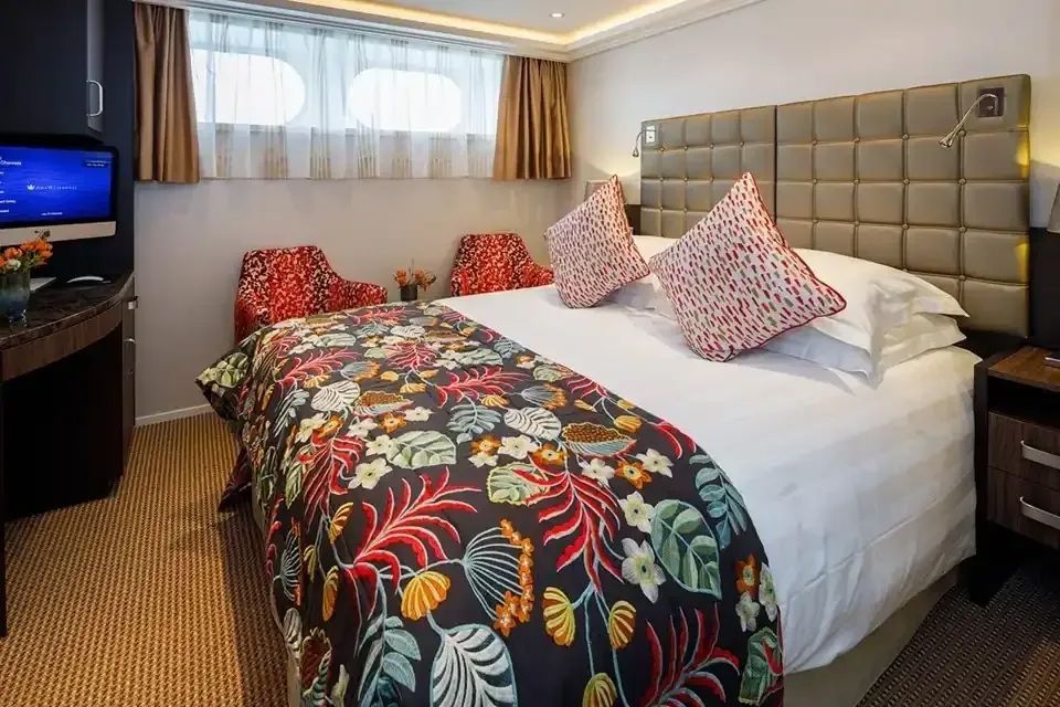 A hotel room with a queen bed featuring a tropical floral runner, two red patterned chairs, and a window with curtains.