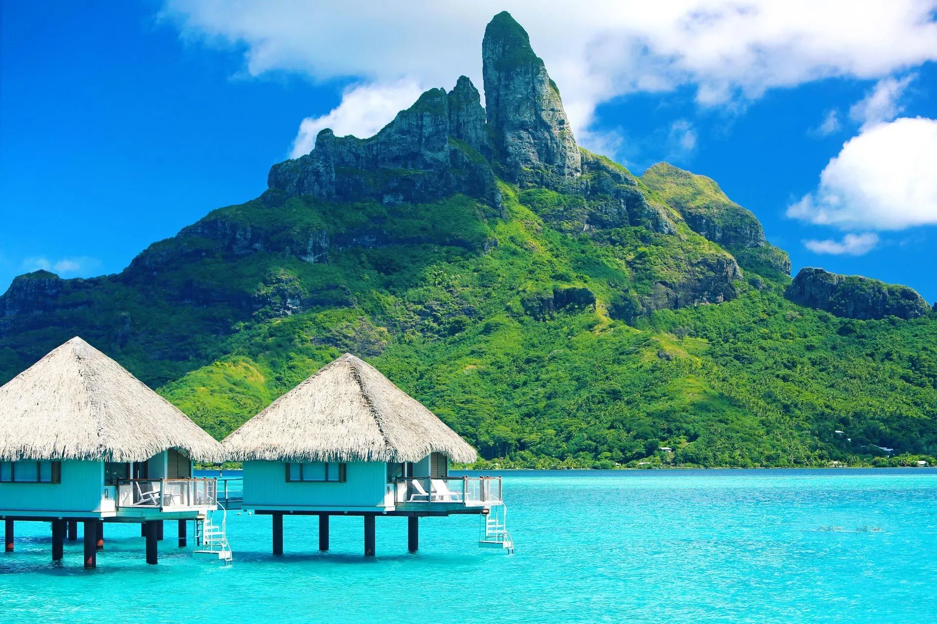 Overwater bungalows in turquoise water with a lush green mountain backdrop.