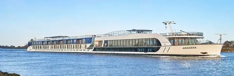 A long, white river cruise ship with large windows sails on calm blue water under a clear sky.