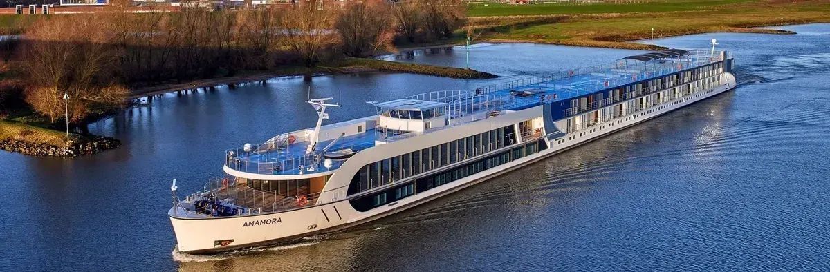 A sleek white river cruise ship travels along a calm, blue river near a tree-lined bank during the day.