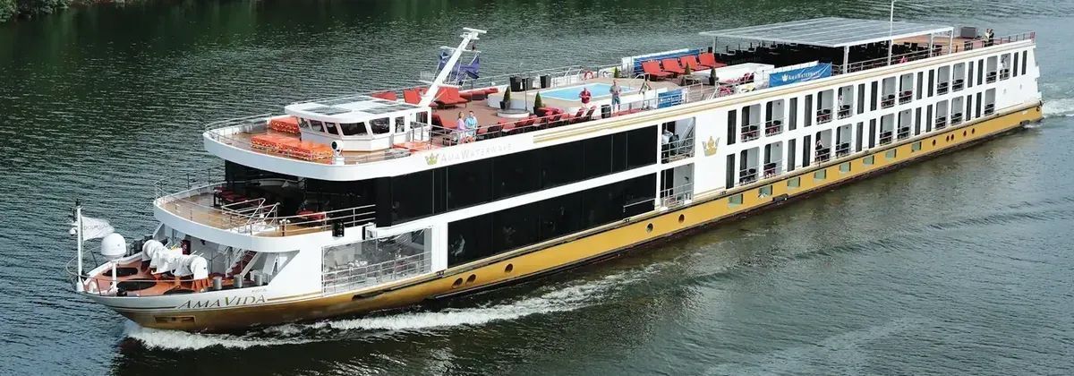A white and gold river cruise ship sailing on a dark body of water.