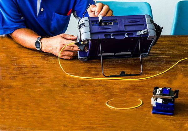 A man is sitting at a wooden table using a device to test a fiber optic cable.