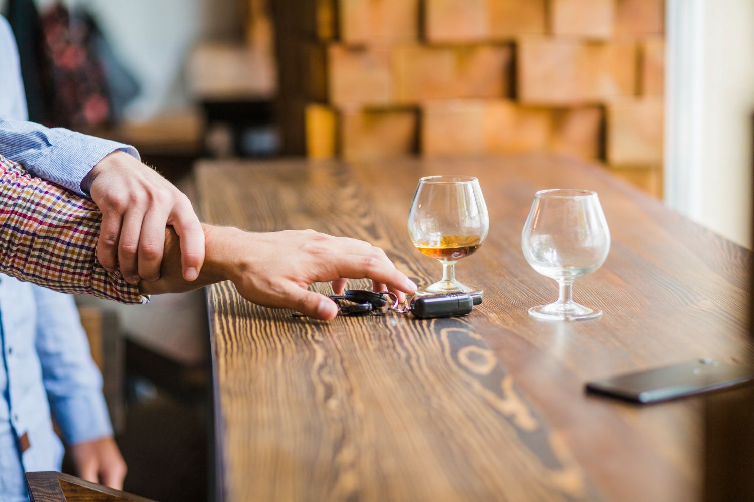 A person's hand reaches for car keys on a bar, another person's hand restrains. Two glasses of liquid and phone on the bar.