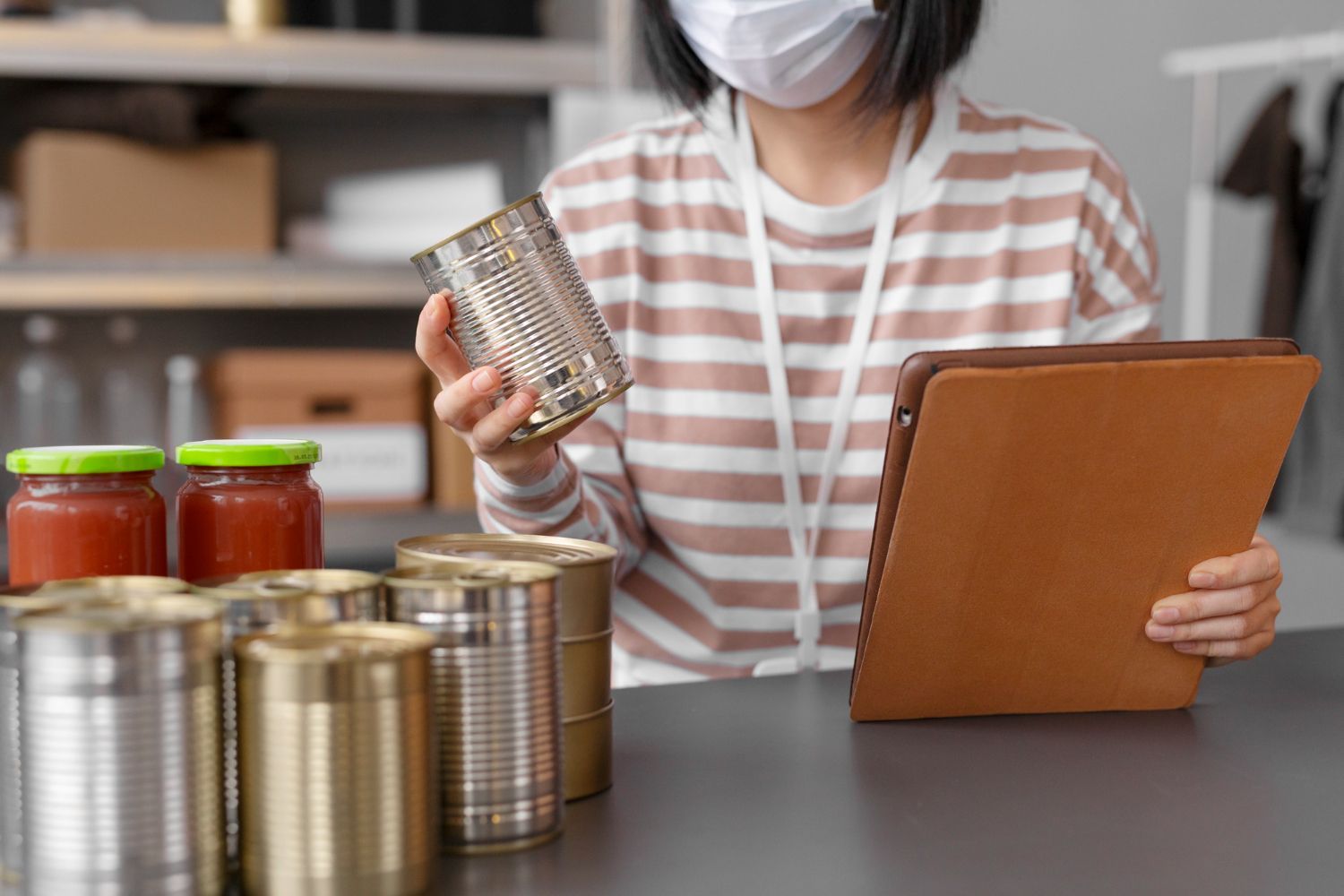 Person wearing a mask holding a can, looking at a tablet, surrounded by canned goods.