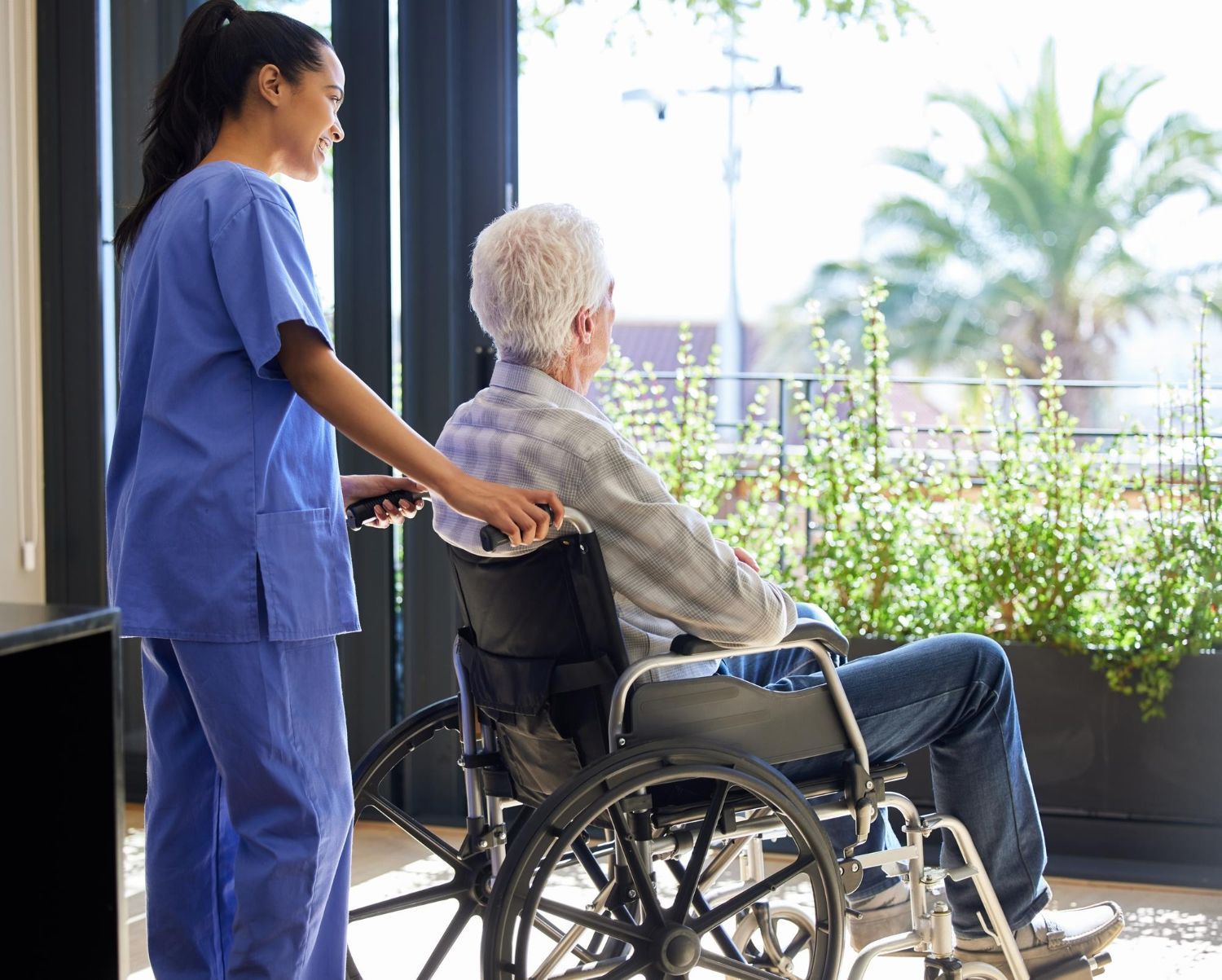 Caregiver pushing a person in a wheelchair near a window with greenery.