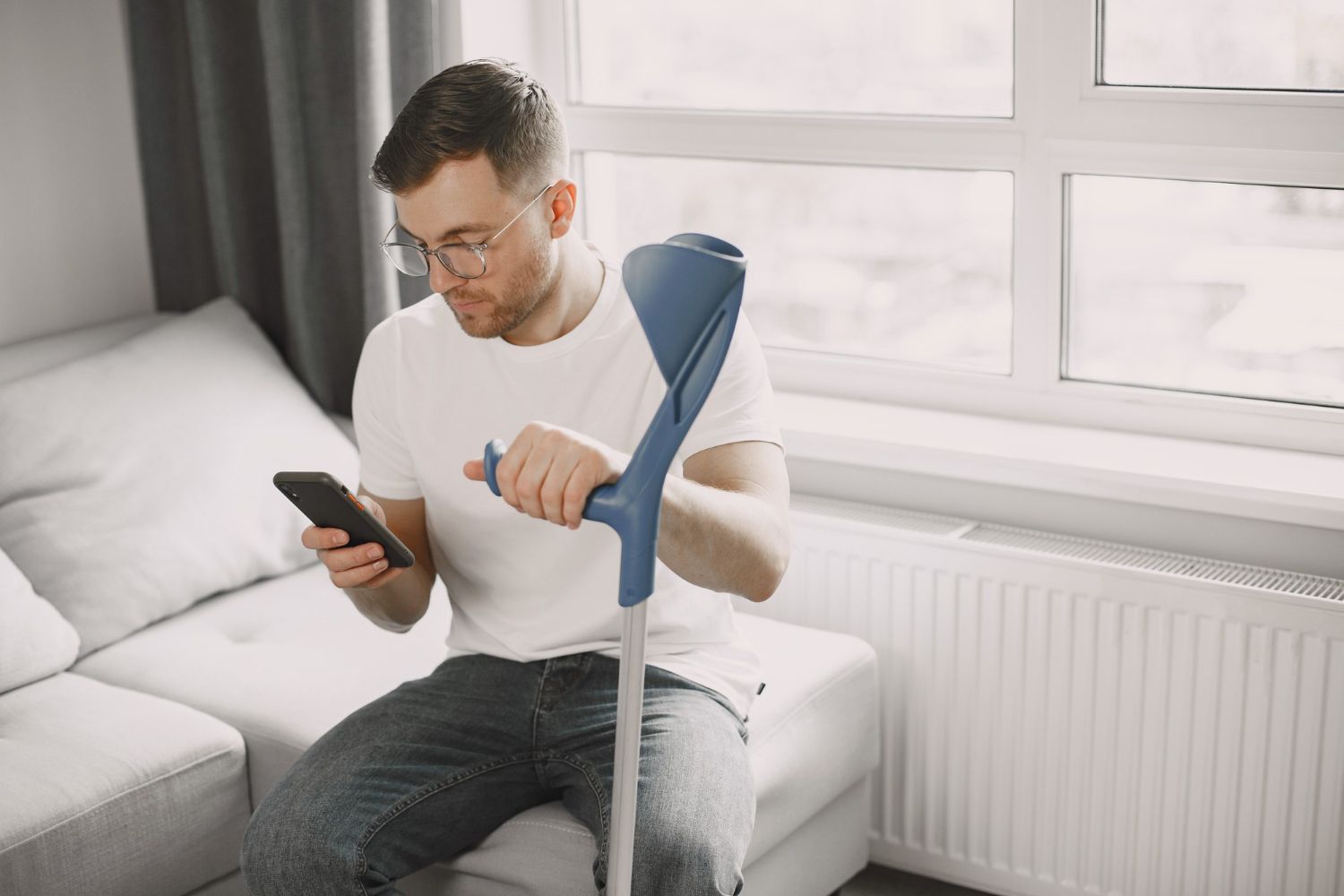 Man with eyeglasses and crutch looking at phone on a couch near a window.