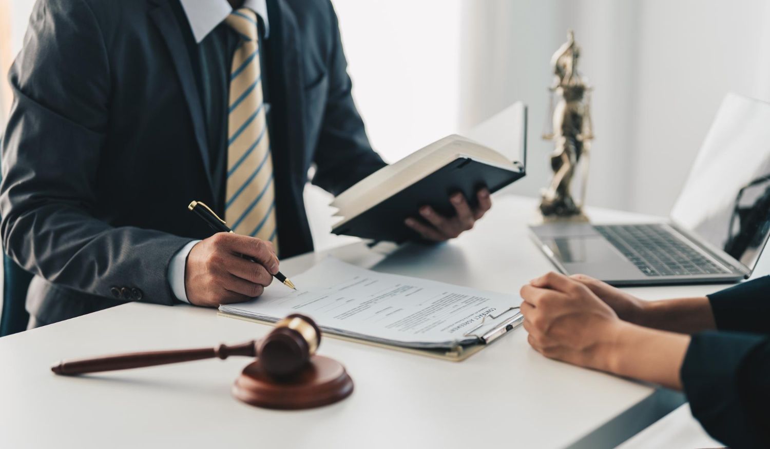 Lawyer writing on documents with a client; gavel, book, and laptop on table.