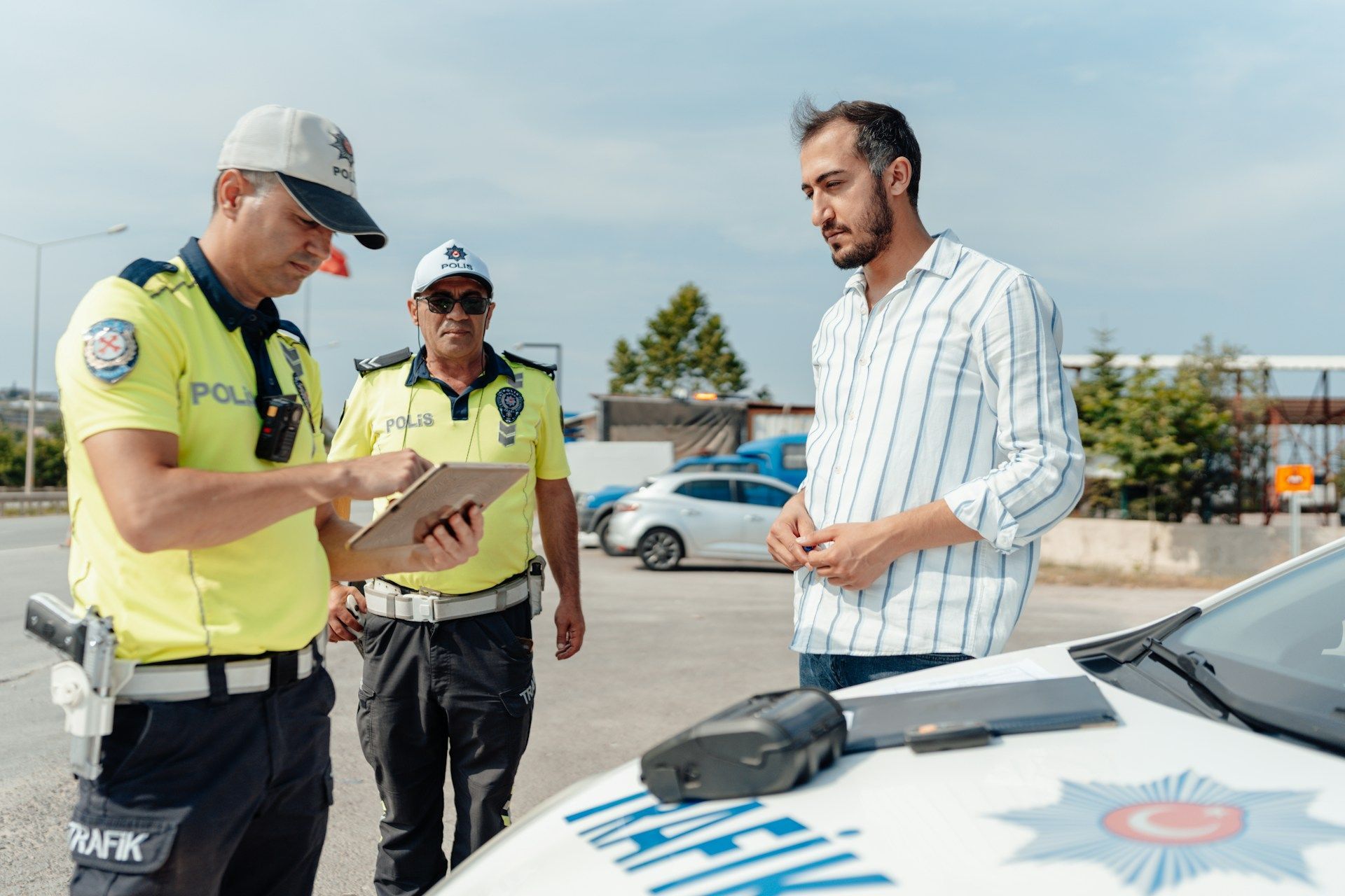 Two Turkish traffic police officers reviewing a tablet with a driver next to a police car in daylight.