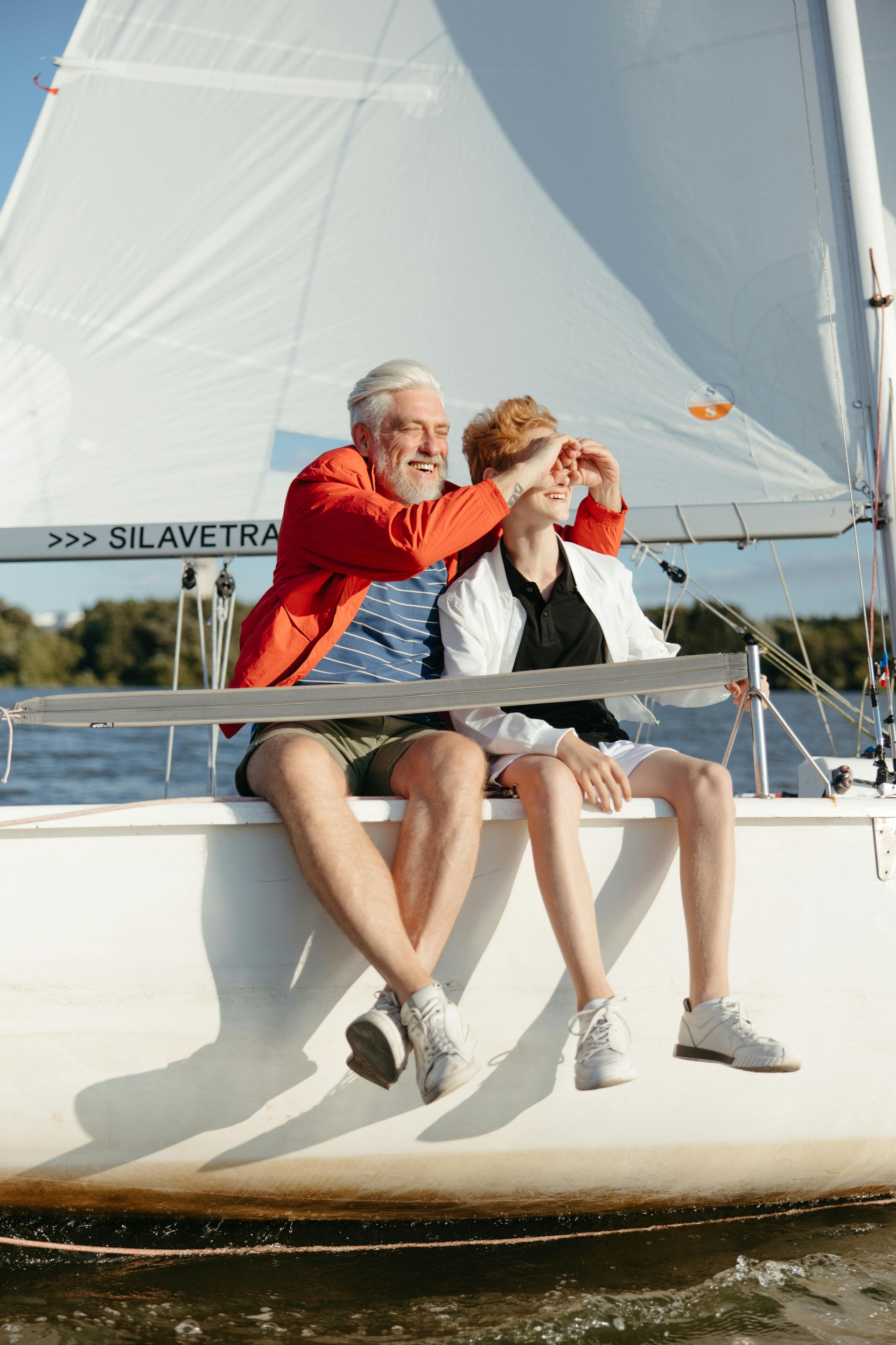Man covering a person's eyes on a sailboat. They're sitting outside, smiling. The person is wearing a white jacket.