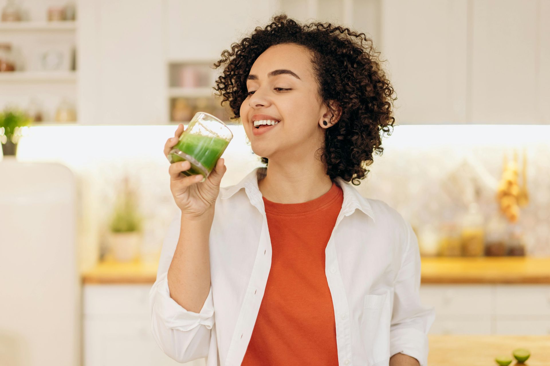 A woman is drinking a green smoothie in a kitchen.