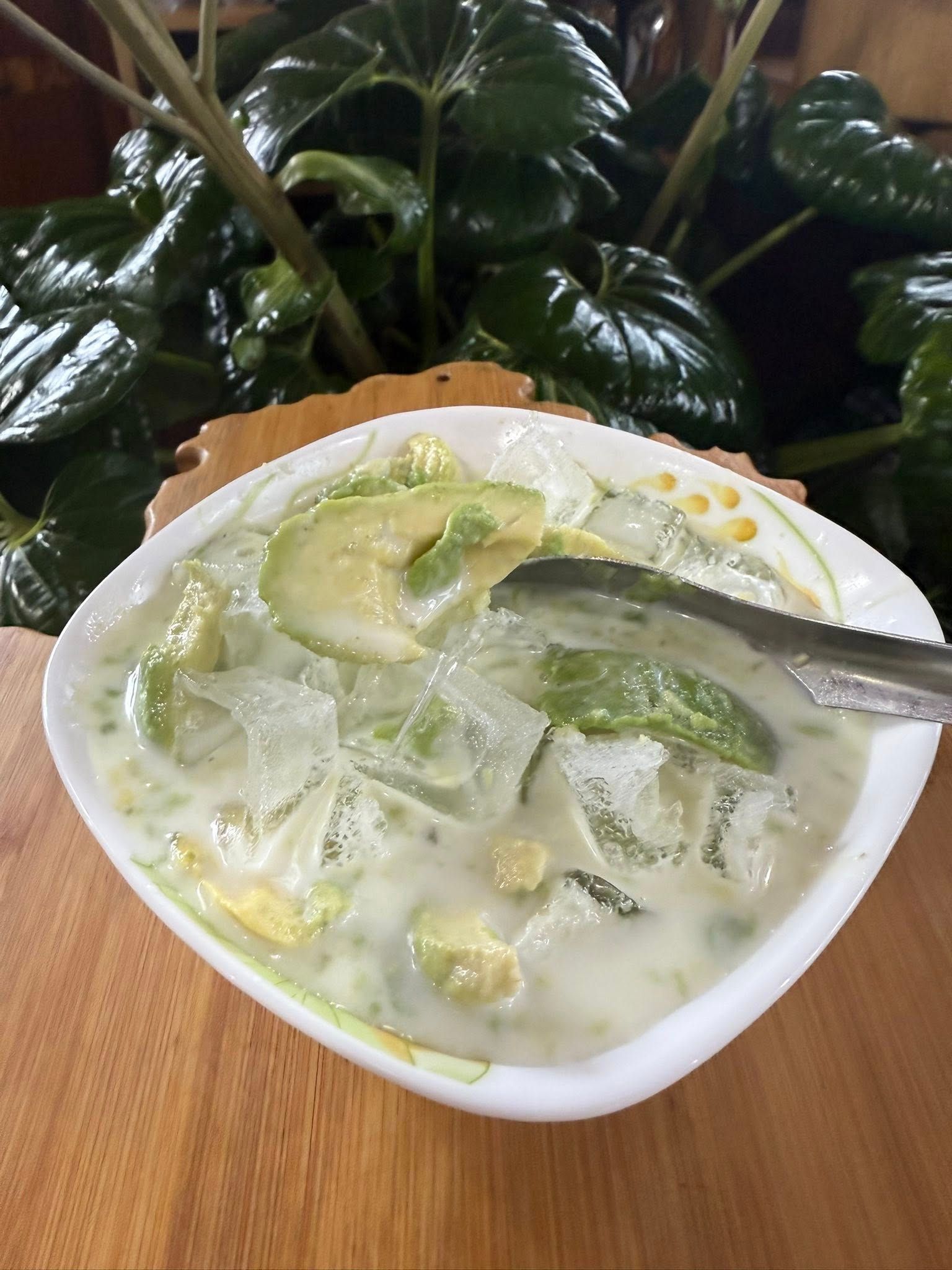 Bowl of avocado dessert with ice, cream, and green pieces. On a wooden table.