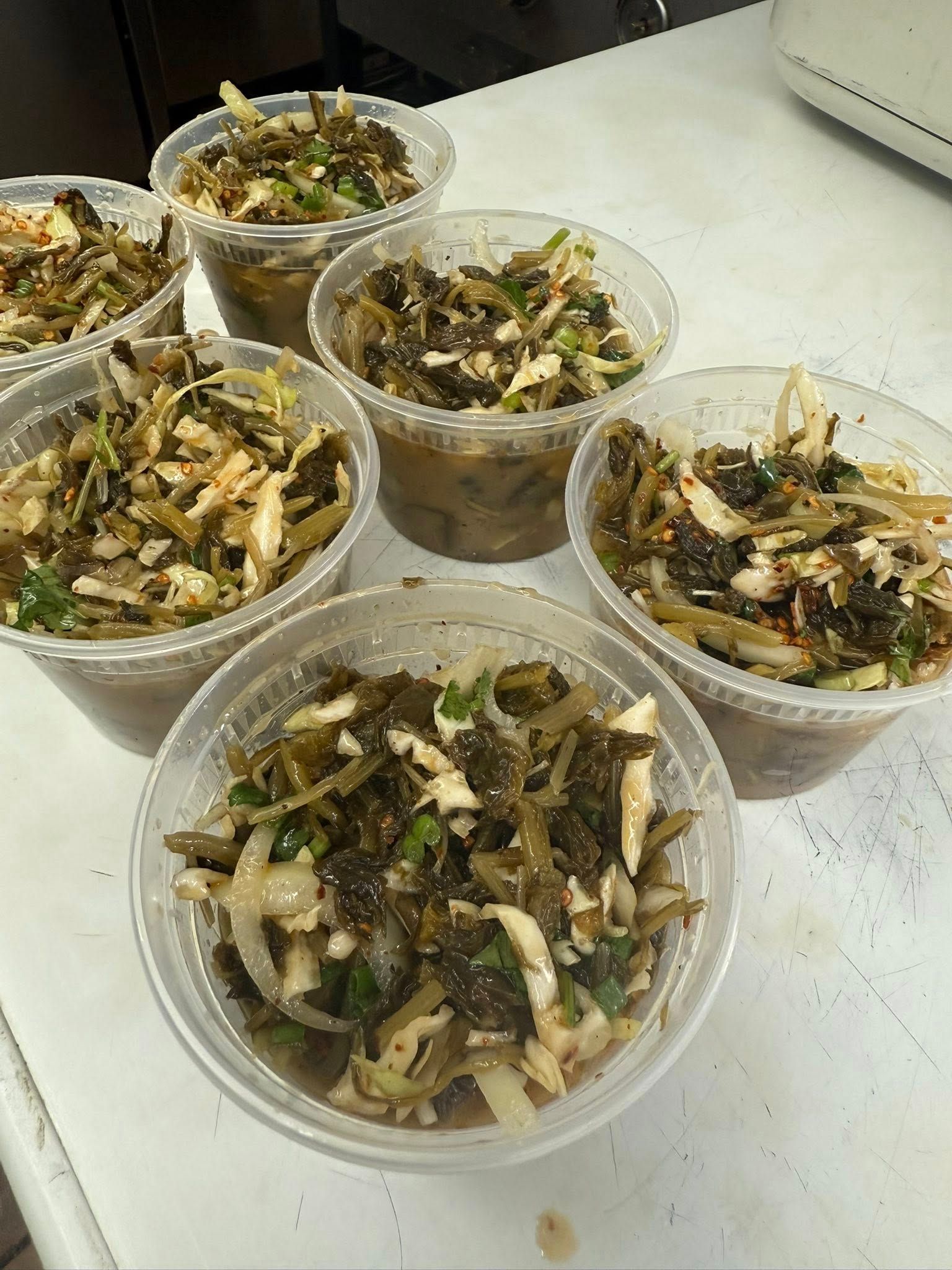 Small containers of a salad with dark green seaweed, white vegetables, and a dark sauce on a white surface.