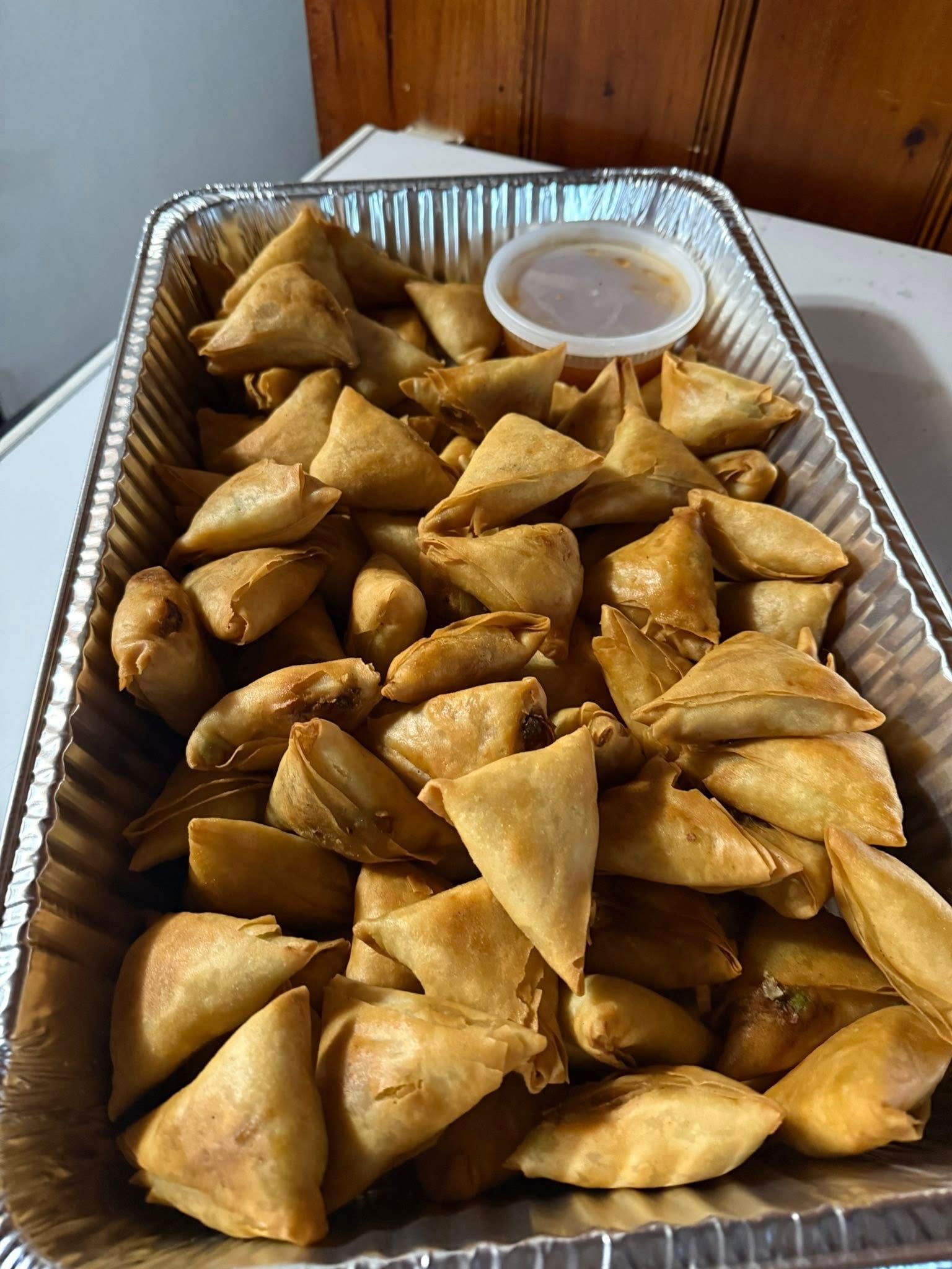 Tray of golden-brown samosas with dipping sauce in a small container.