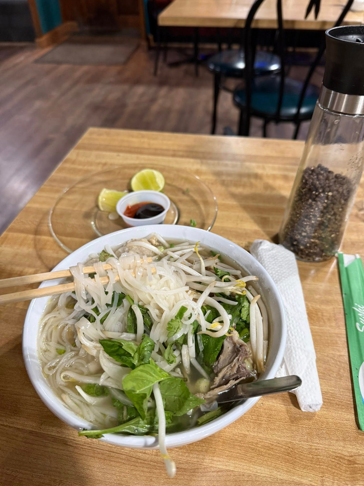 Bowl of pho with chopsticks, lime, sauce, pepper, and napkin on a wooden table in a restaurant.