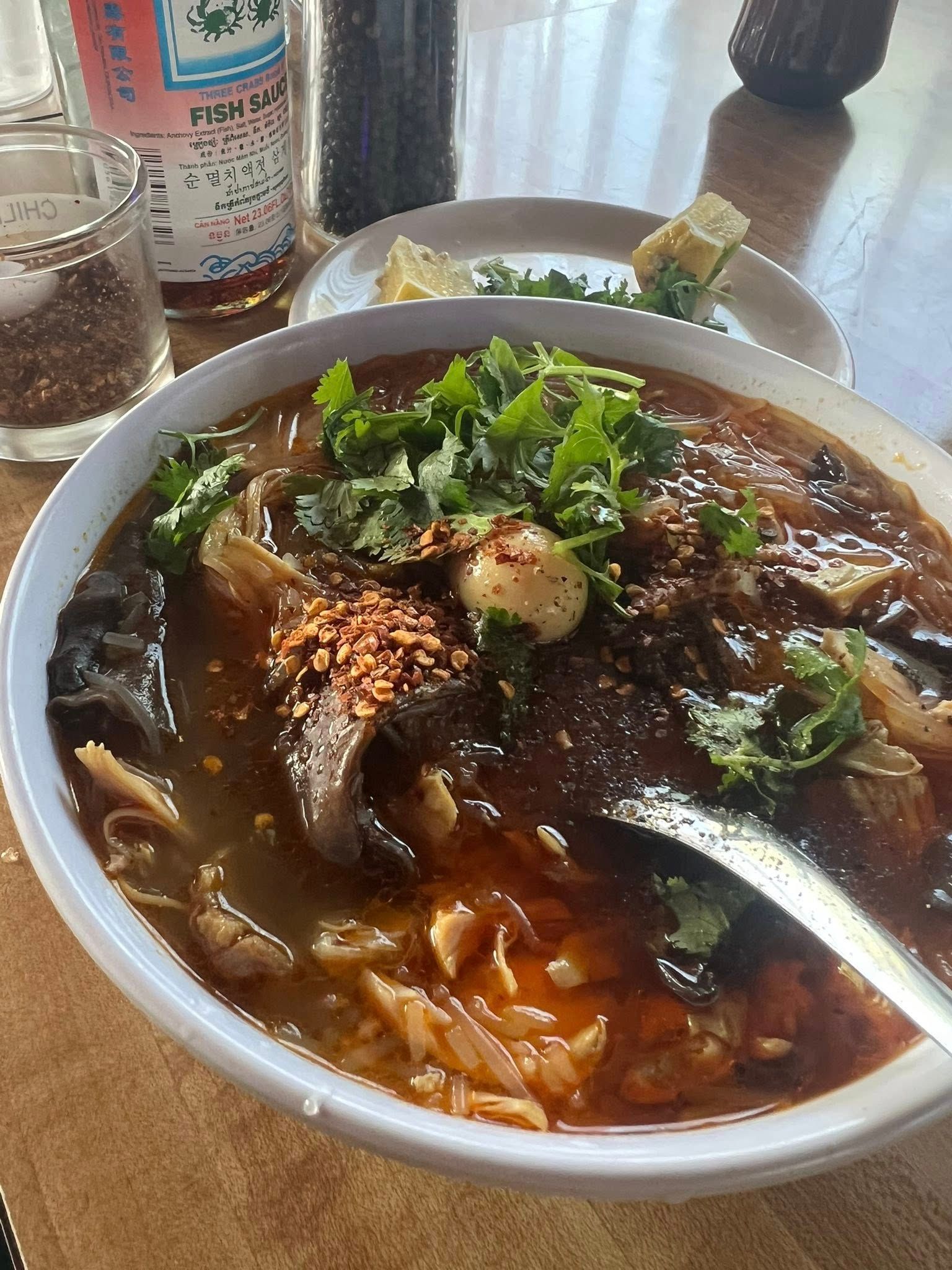 Bowl of reddish-brown soup with cilantro, meat, and a spoon. Side dishes and bottles in the background.