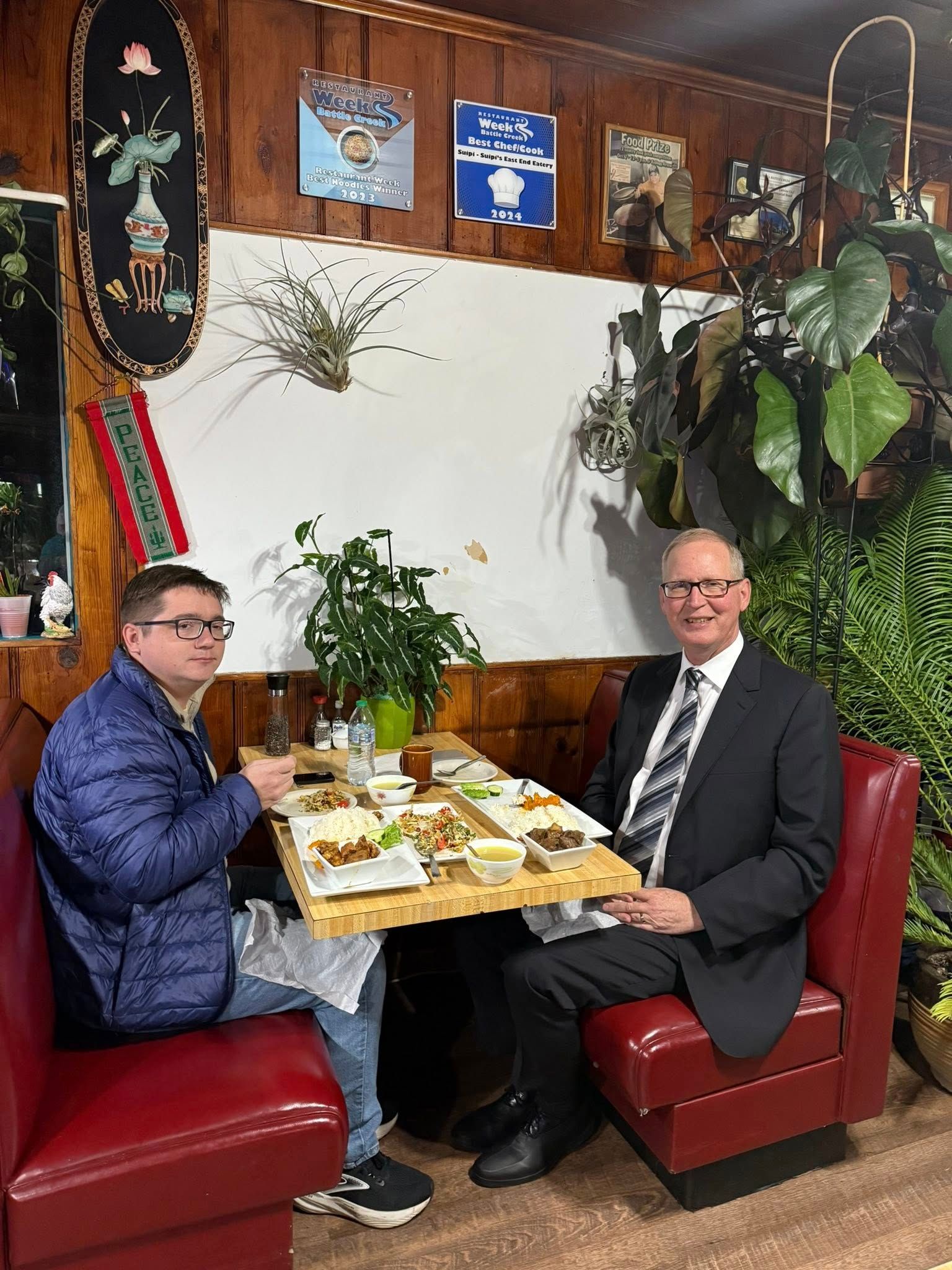 Two men seated at a restaurant table. One in a suit, the other in a blue jacket. Plates of food on the table.