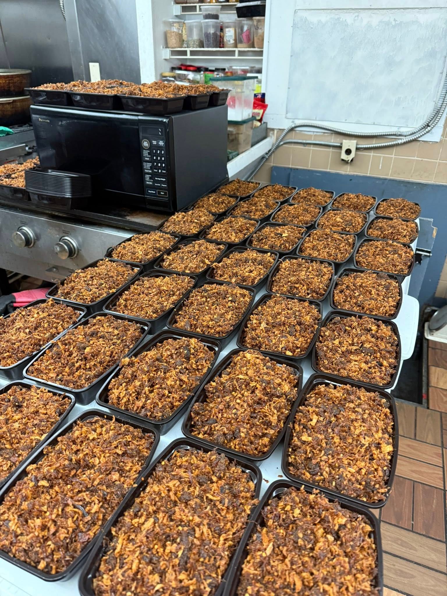 Many trays filled with food, likely for meal prep, arranged on a countertop near a microwave and a stove.