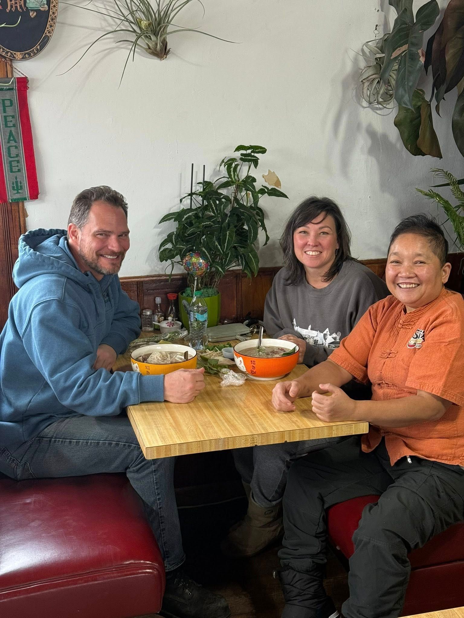 Three people seated at a table in a restaurant. They are smiling. Bowls of food are on the table.
