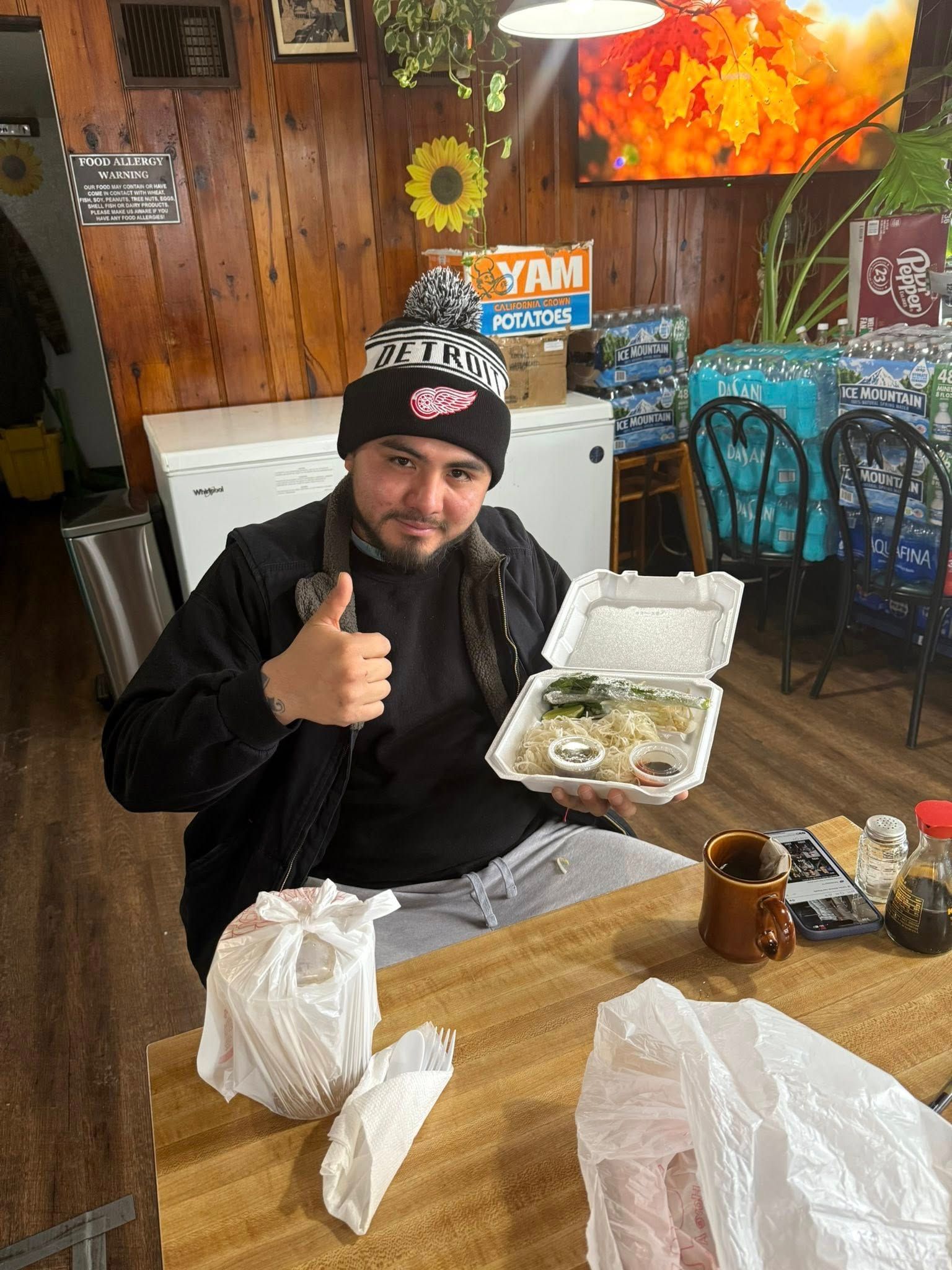 Man with a black and white hat, holding takeout food, giving a thumbs-up inside a restaurant.
