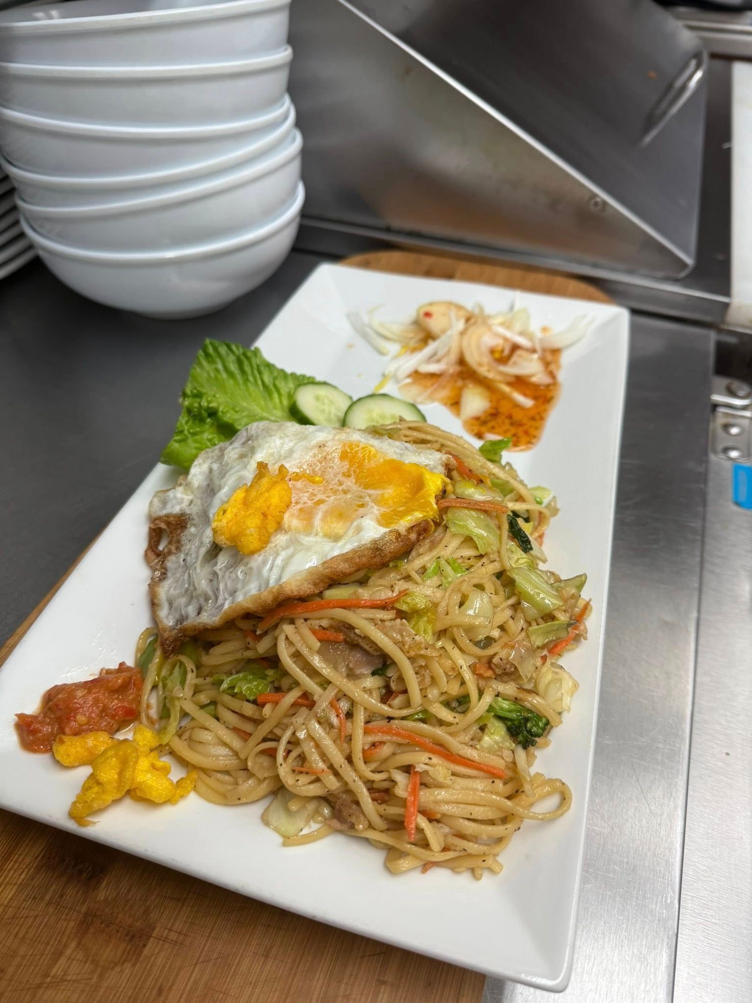 Noodle dish with fried egg, vegetables, and side dishes on a white rectangular plate.