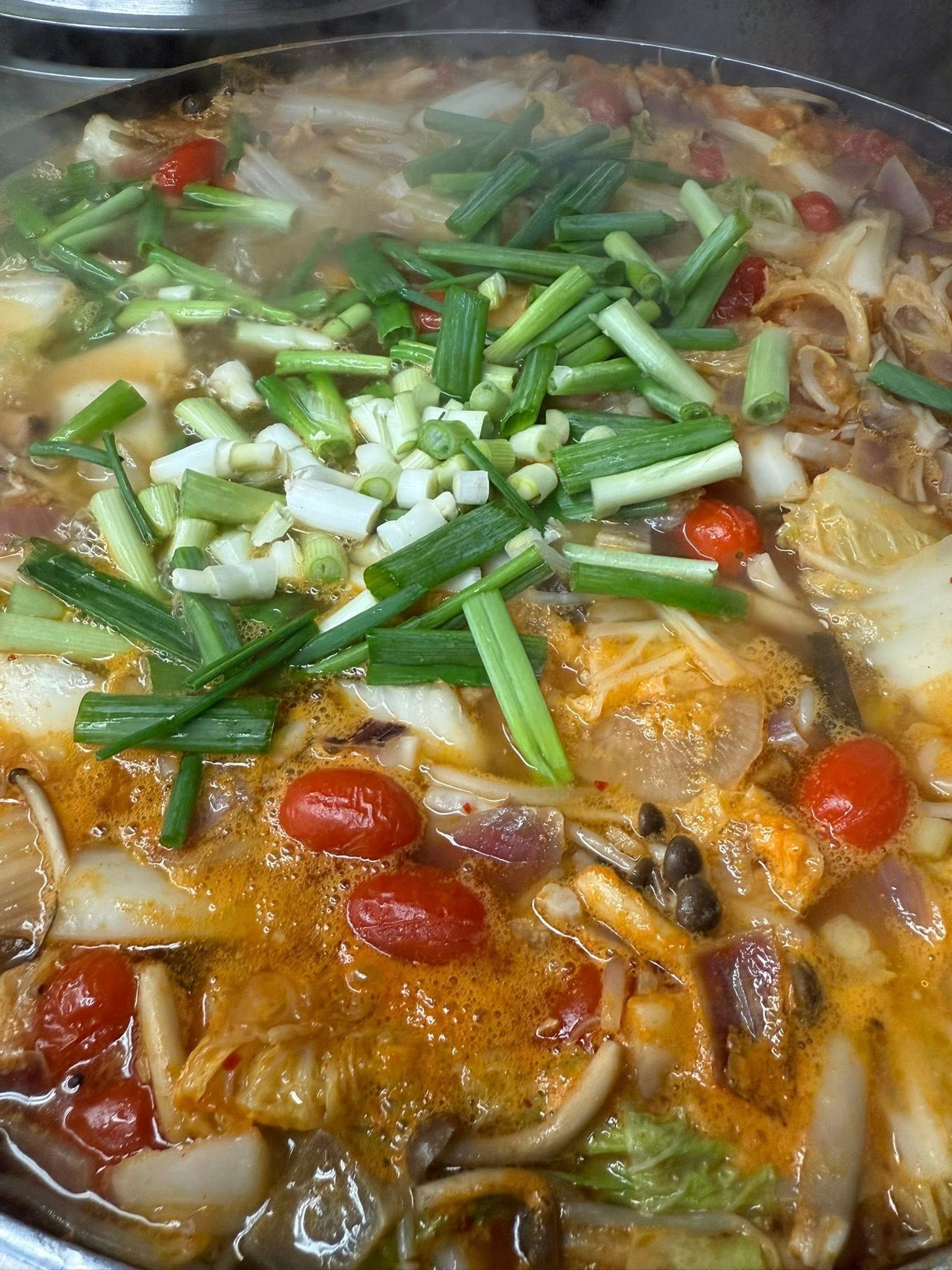 Steaming pot of soup with green onions, cherry tomatoes, and various vegetables.