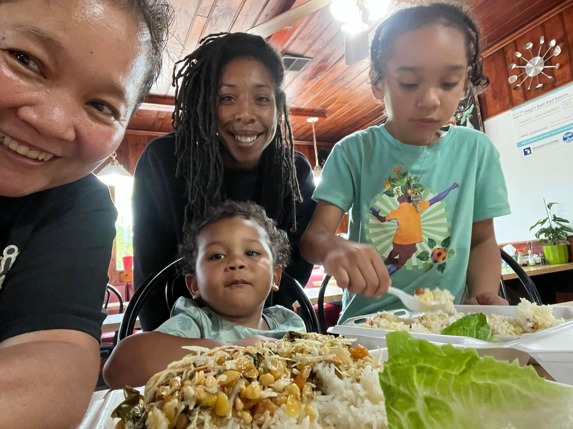 Four people at a table, smiling with plates of food. One person takes a bite, one looks at the camera. Wooden interior.