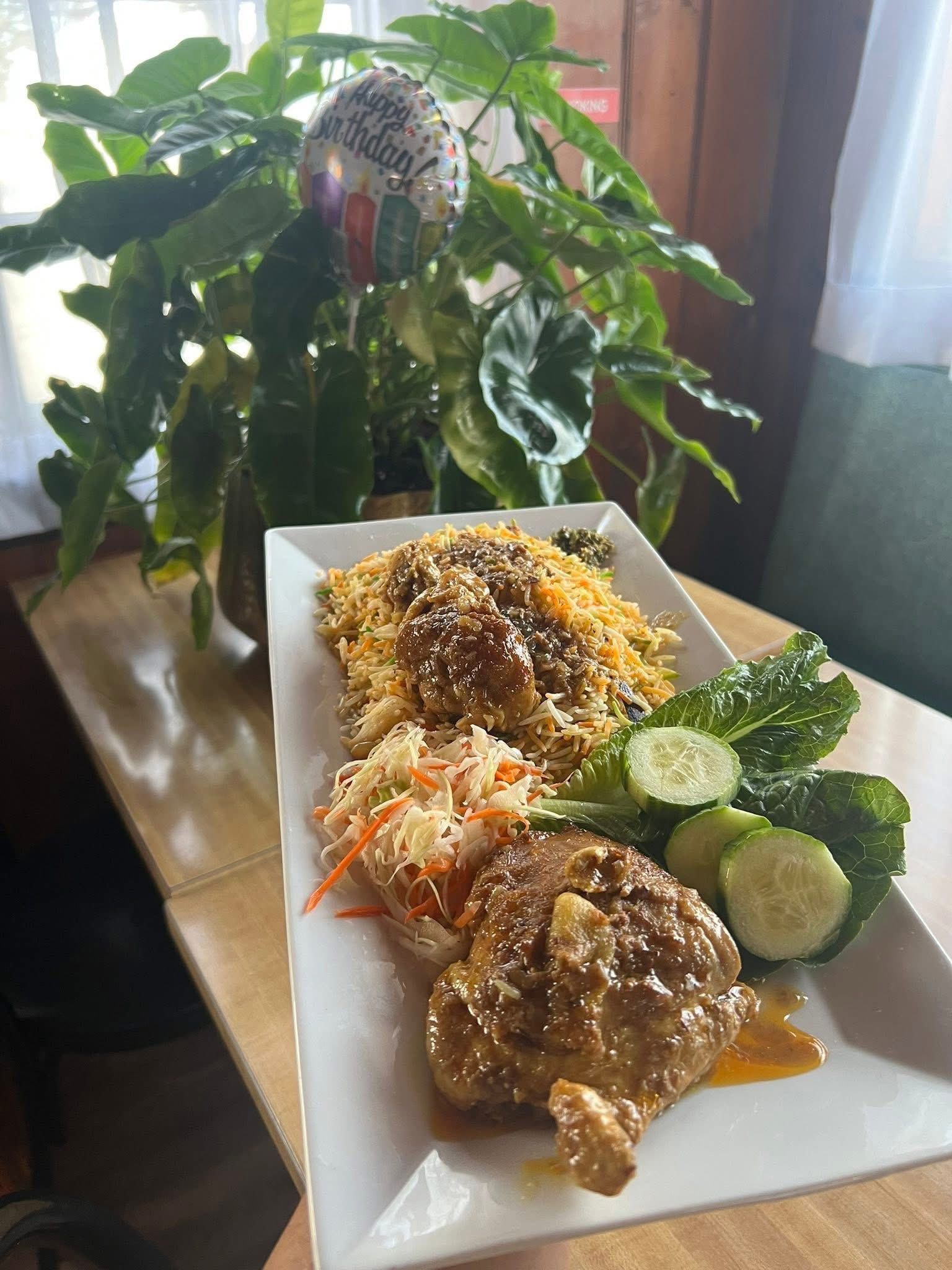 Plate of food: chicken, rice, slaw, cucumbers, with a plant in background.