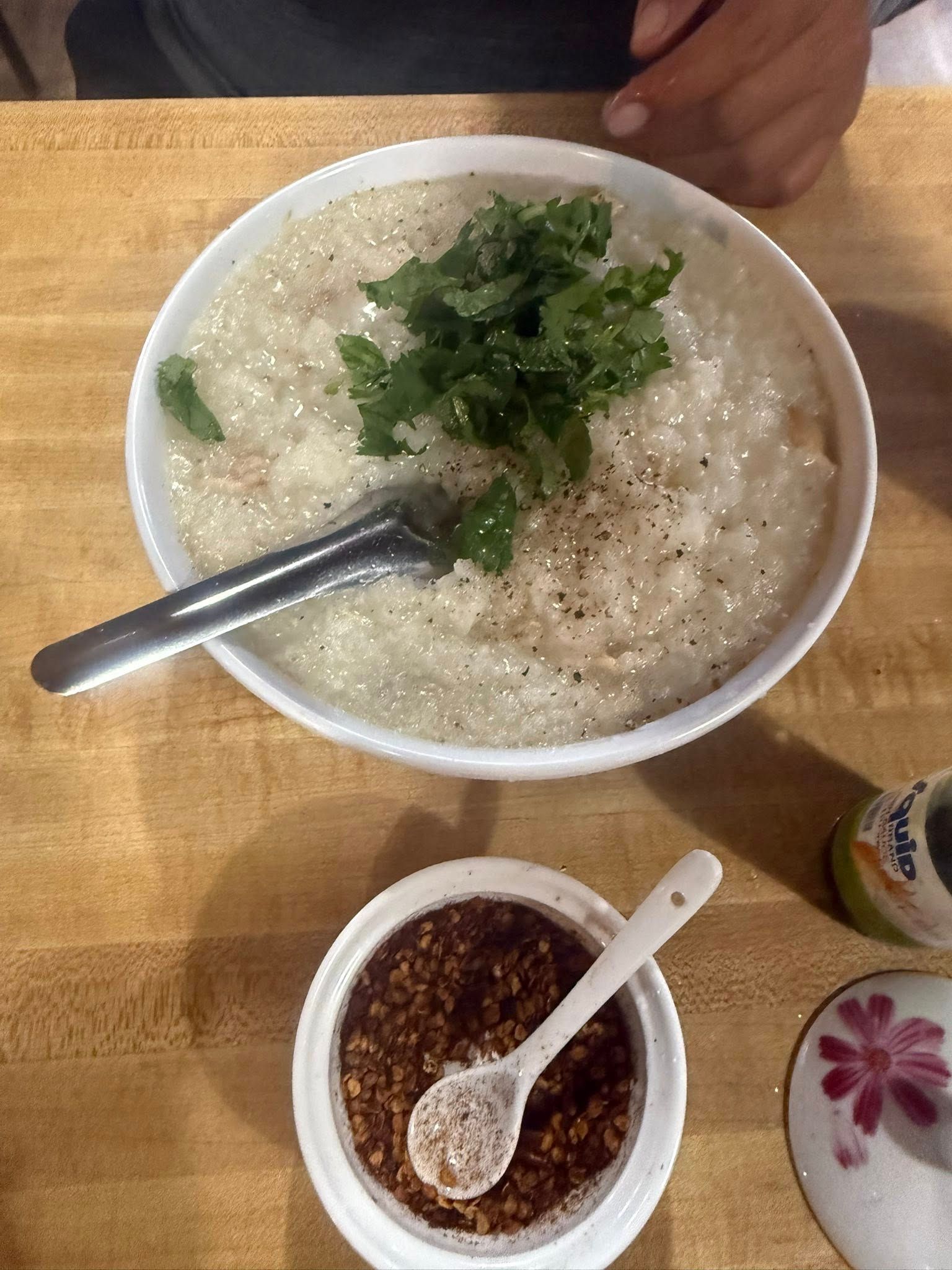 A bowl of congee with herbs, a bowl of chili flakes, and a spoon, on a table.