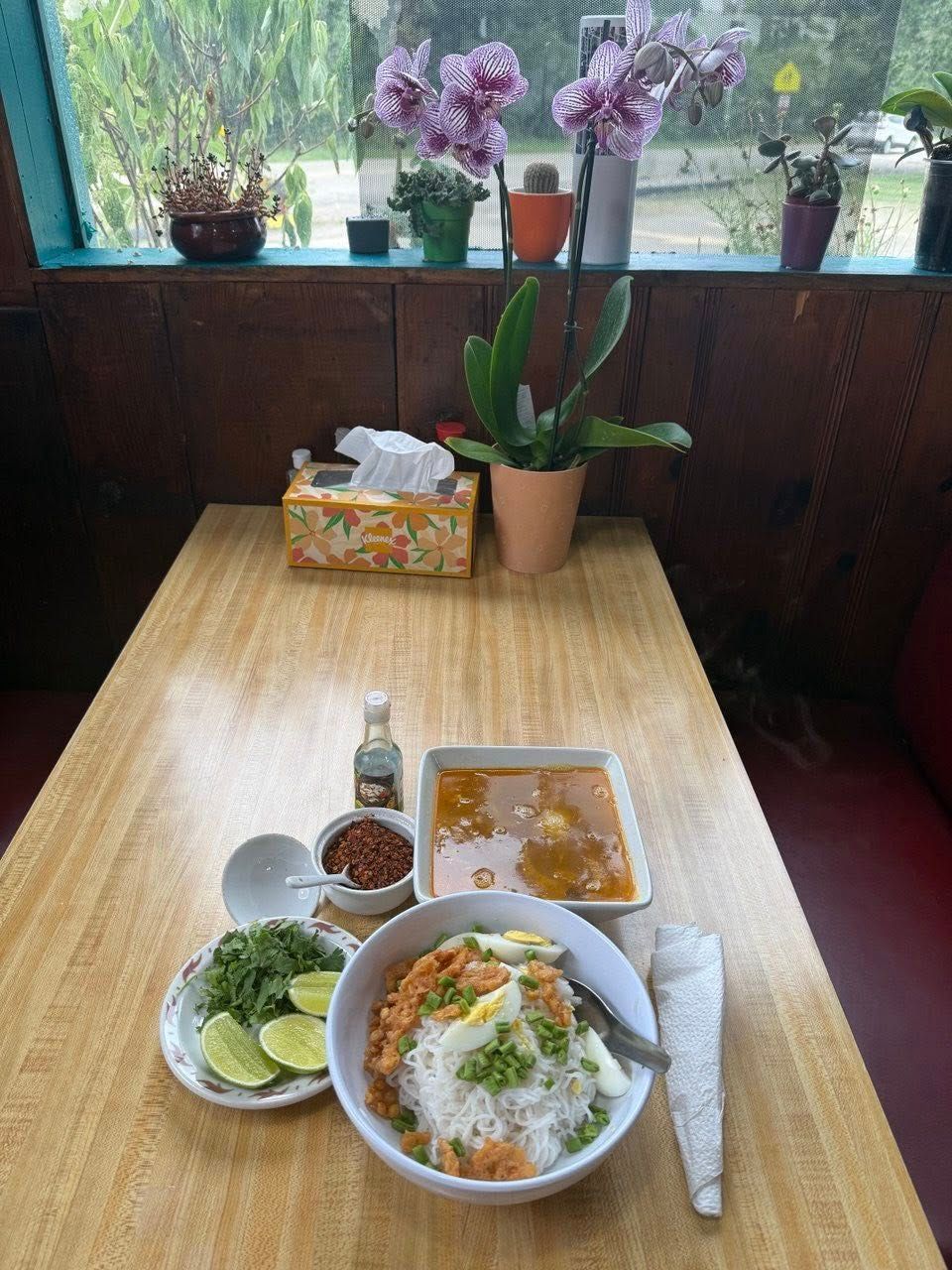Table set with Burmese food, herbs, and condiments. Flowers on windowsill above.