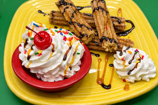 Dessert platter with whipped cream, cherry, churros, and chocolate drizzle on a yellow plate