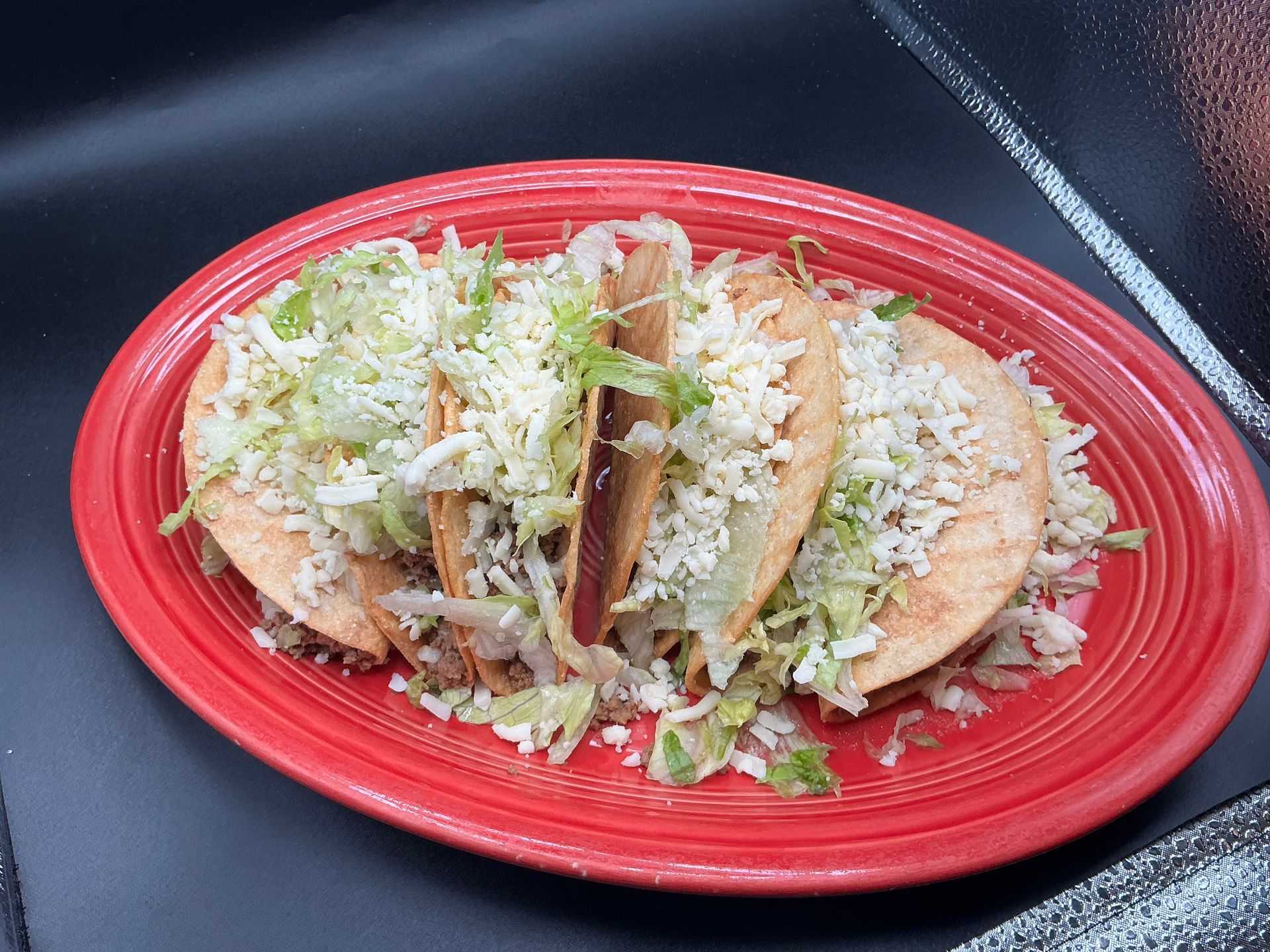 Red plate with three tacos topped with shredded cheese, lettuce, and salsa on a dark table