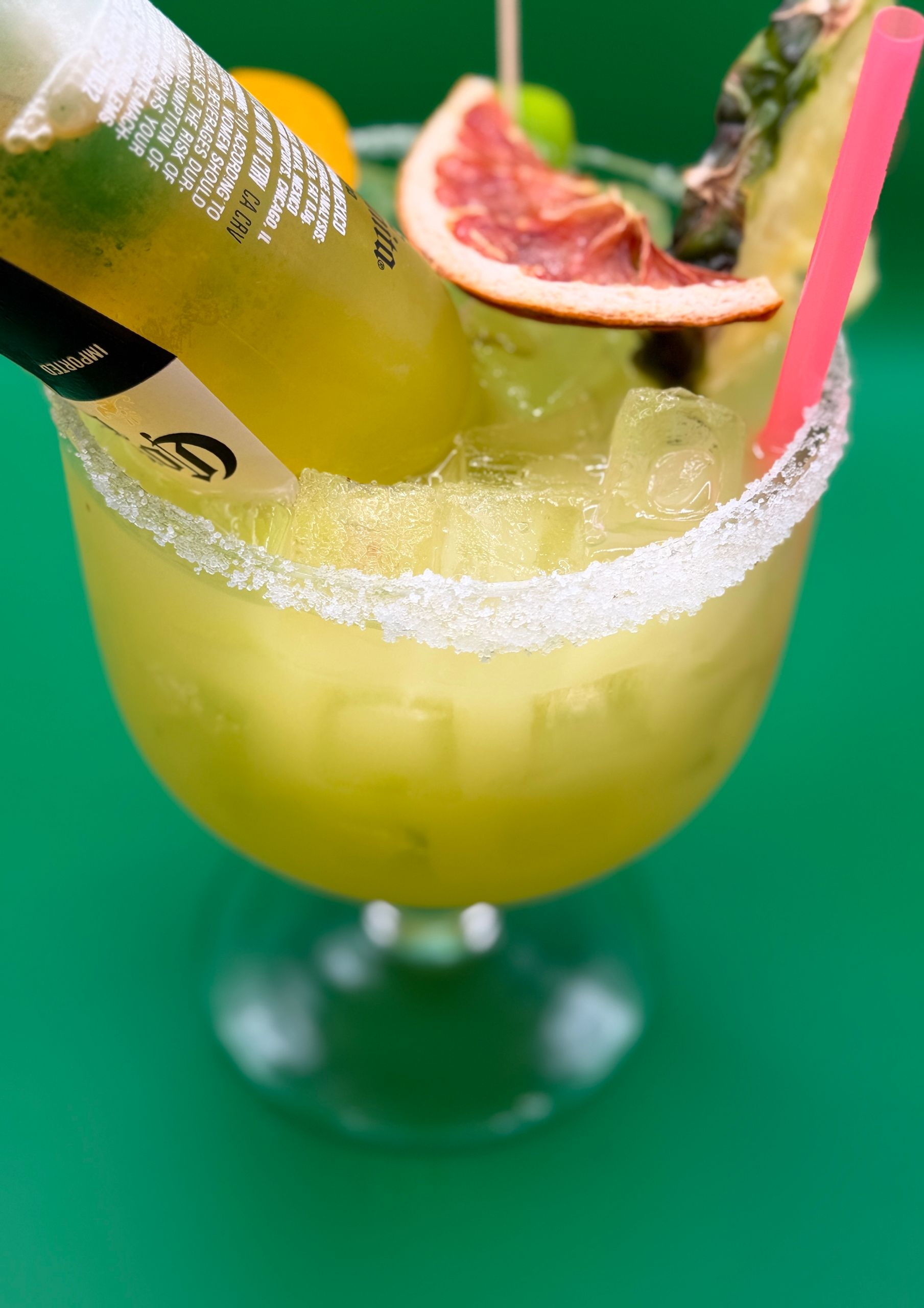Green cocktail with a salted rim, pink straw, and grapefruit garnish in a stemmed glass