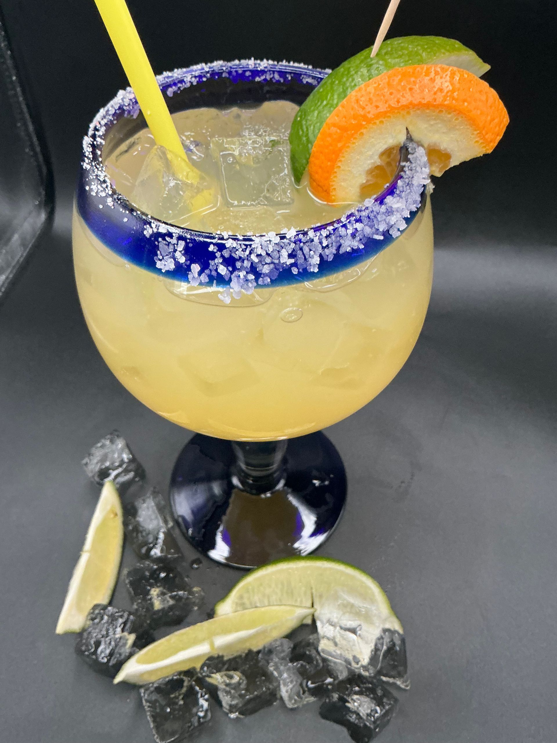 Yellow cocktail with blue-sugar rim and citrus garnish in a stemmed glass, with lime wedges below