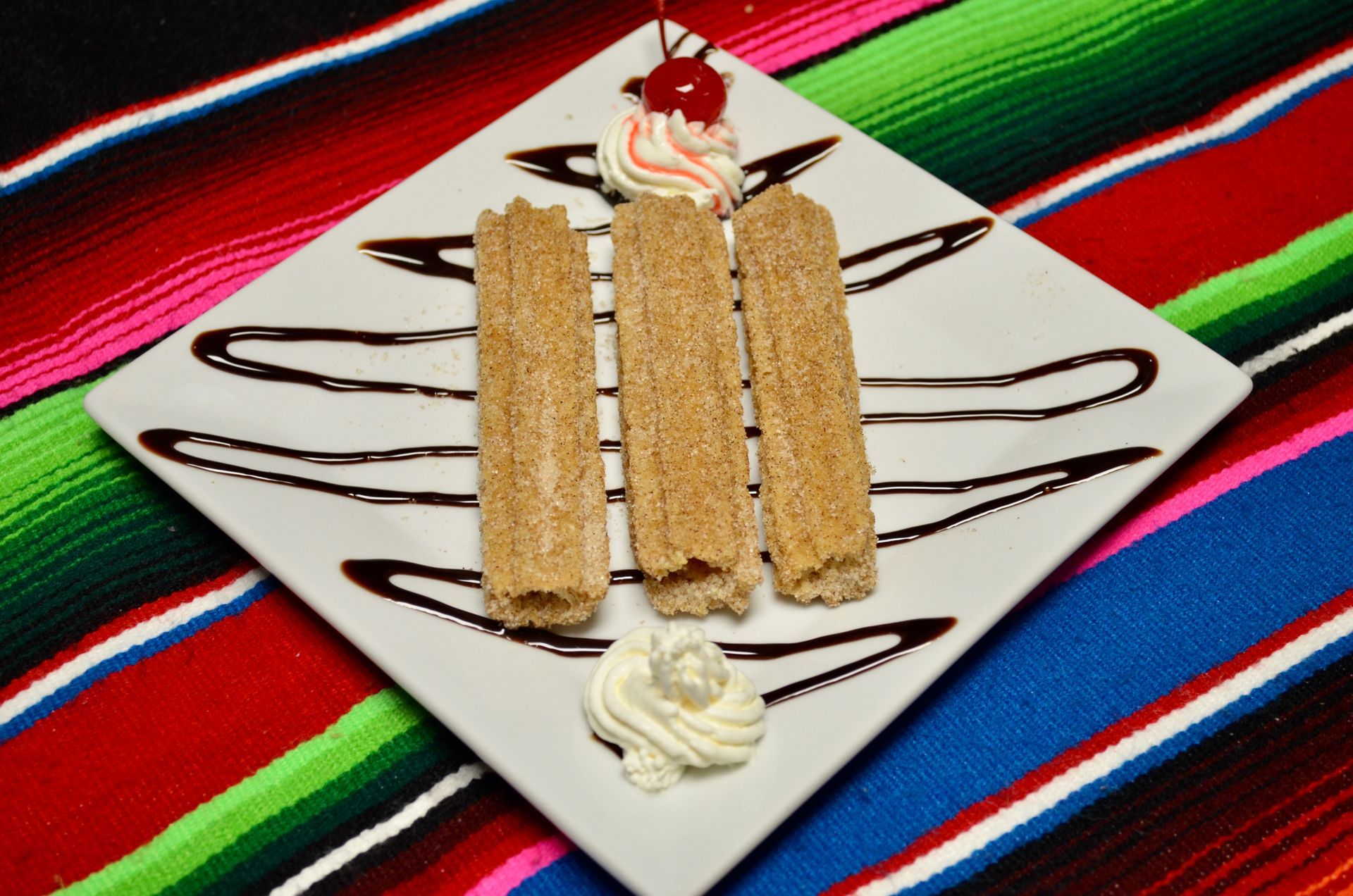 Churros with cinnamon sugar, drizzled chocolate, whipped cream, and cherry on a white plate, patterned fabric background.
