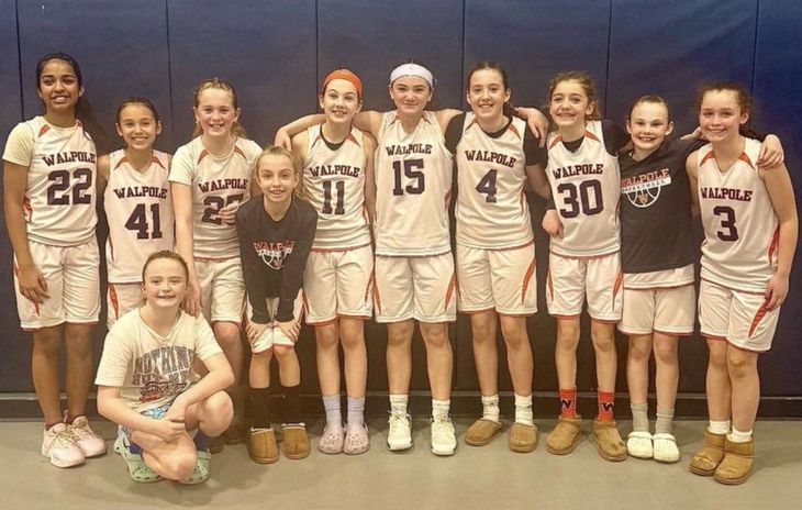 Youth basketball team in white uniforms posing indoors, with one girl kneeling in front and trophies on the floor