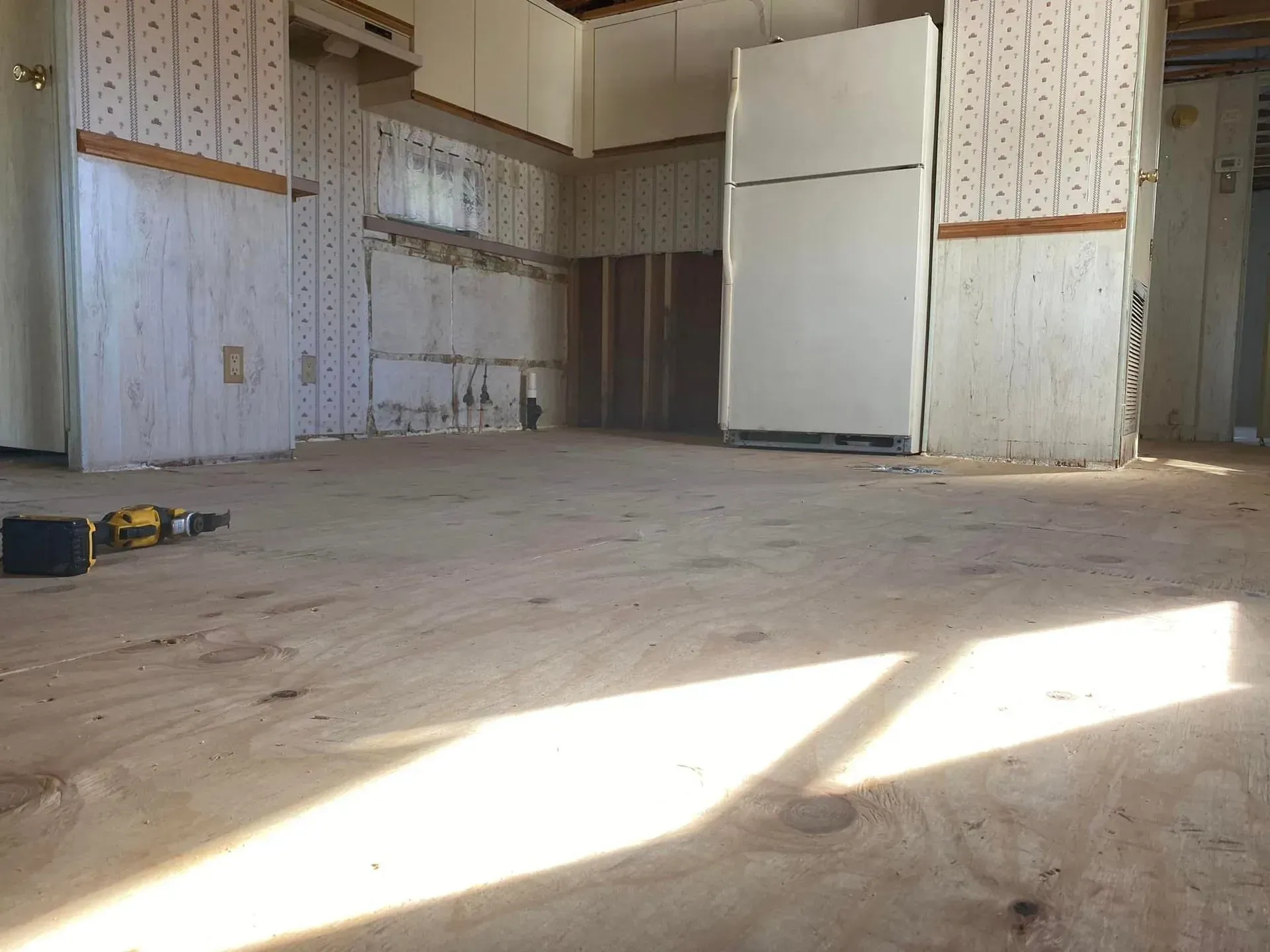 A gutted kitchen with plywood floor, cabinets, and a refrigerator. Sunlight streams across the floor. A gutted kitchen with plywood floor, cabinets, and a refrigerator. Sunlight streams across the floor.