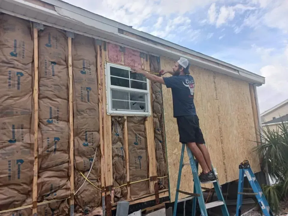 Man on ladder, installing siding on house with exposed insulation and window. Sunny day.