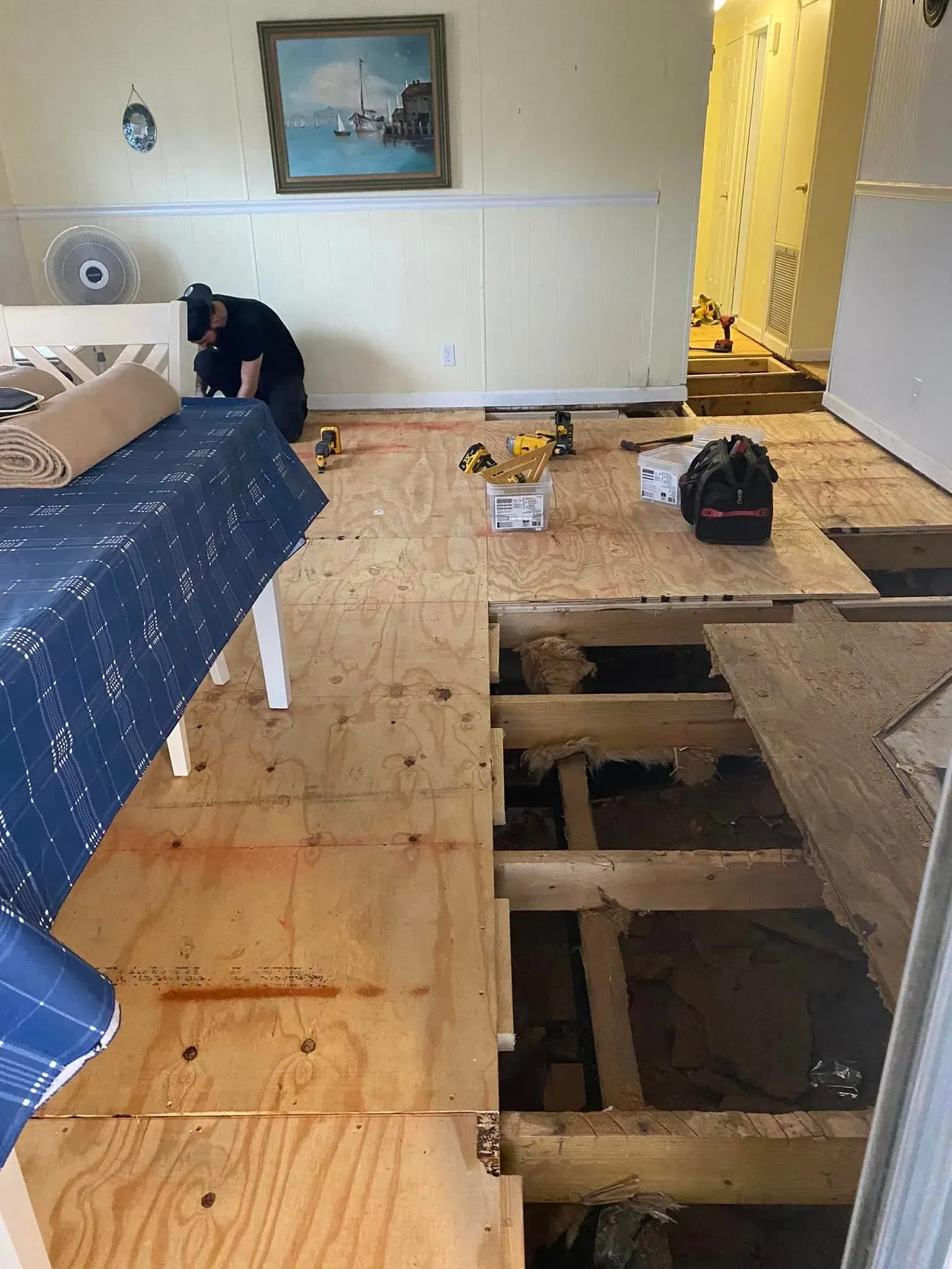 Person working on exposed subfloor in a room; tools, furniture, and painting on walls visible. Person working on exposed subfloor in a room; tools, furniture, and painting on walls visible.